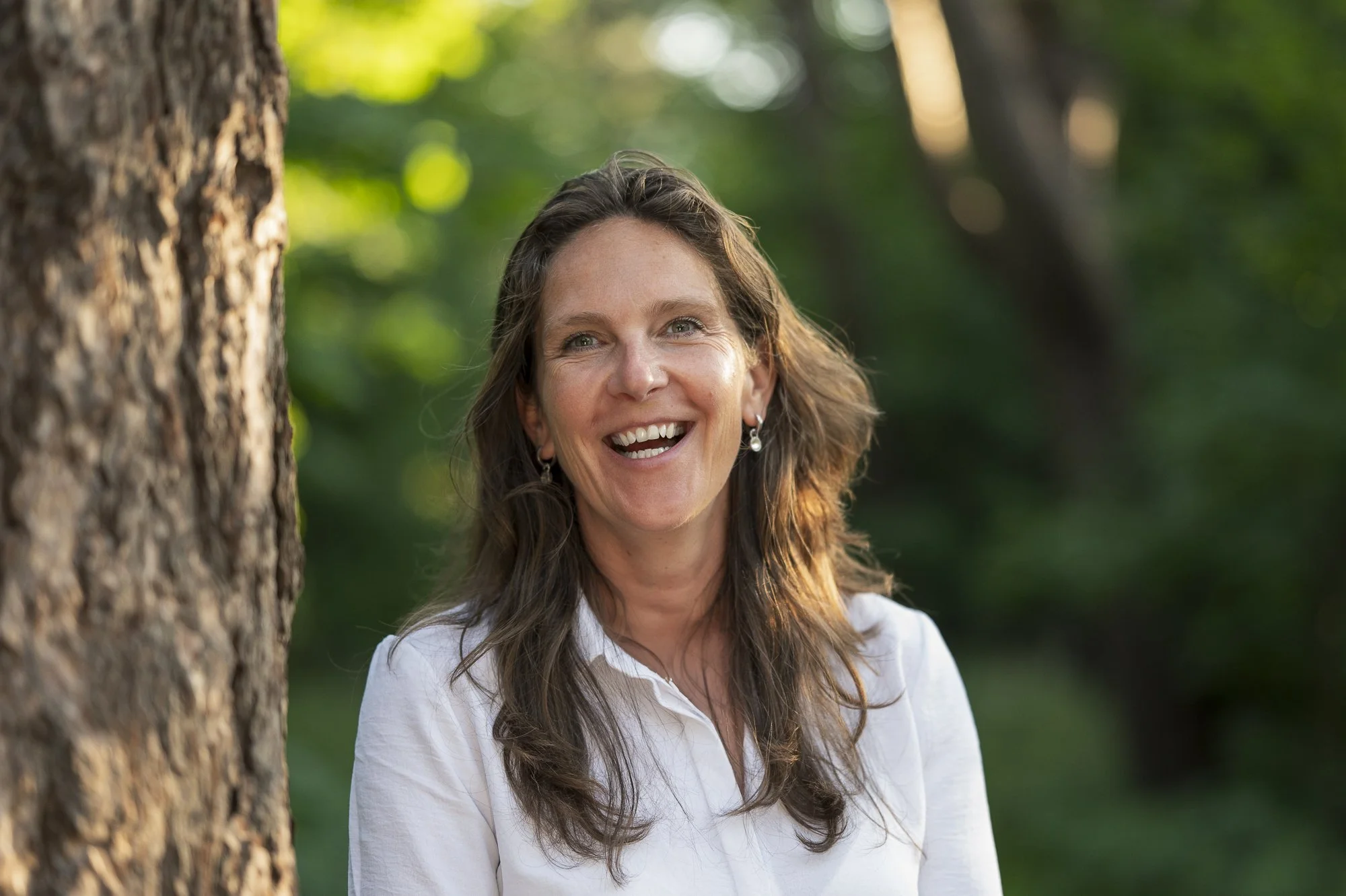 A woman with long, wavy brown hair smiling outdoors with trees and greenery in the background.