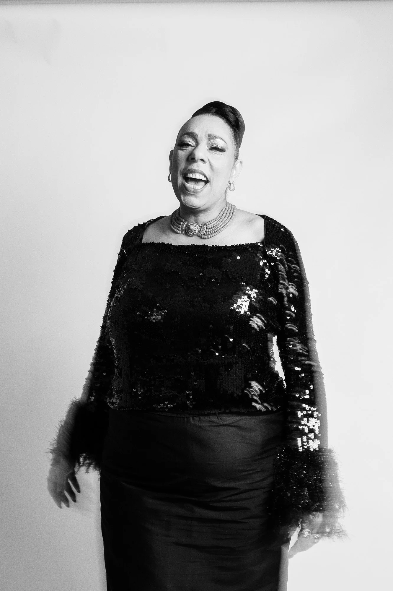A woman singing passionately, wearing a sparkly black top, a necklace, and earrings, with a light-colored background.