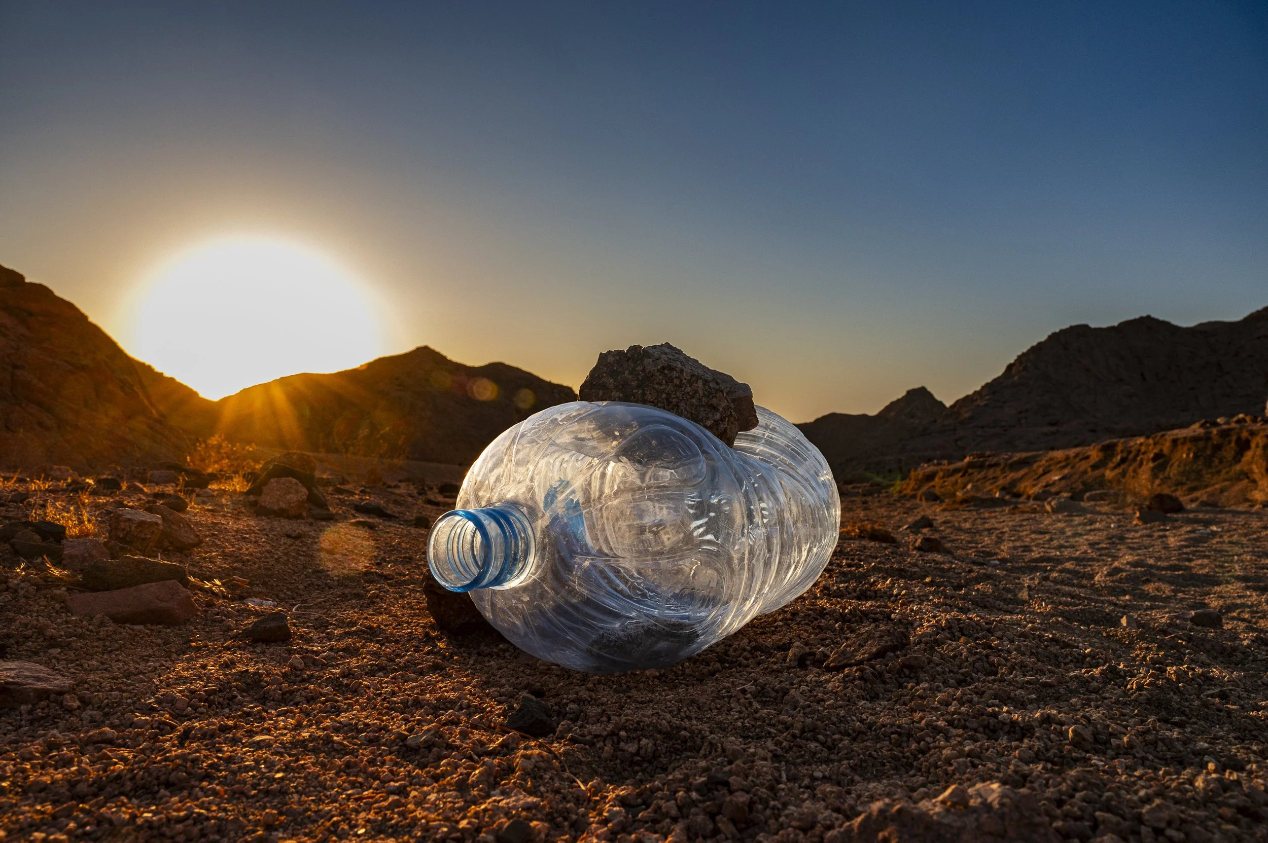 A clear plastic water bottle lying on rocky desert ground with a small rock on top, during sunset in a mountainous landscape.