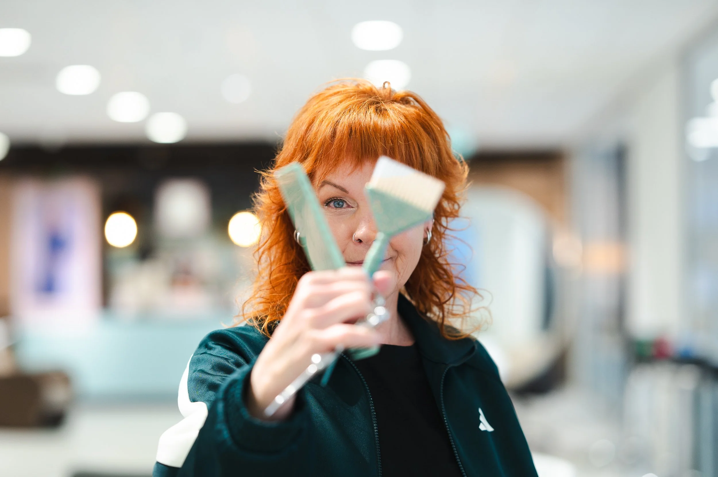 Red-haired woman in a black jacket holding three paintbrushes in front of her face at an indoor art studio or café