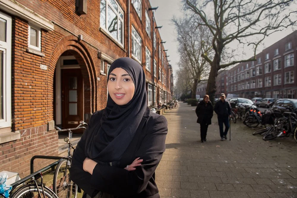A woman wearing a black hijab and black blazer standing on a city sidewalk with her arms crossed, smiling, with brick buildings and bicycles in the background, and two people walking in the distance.