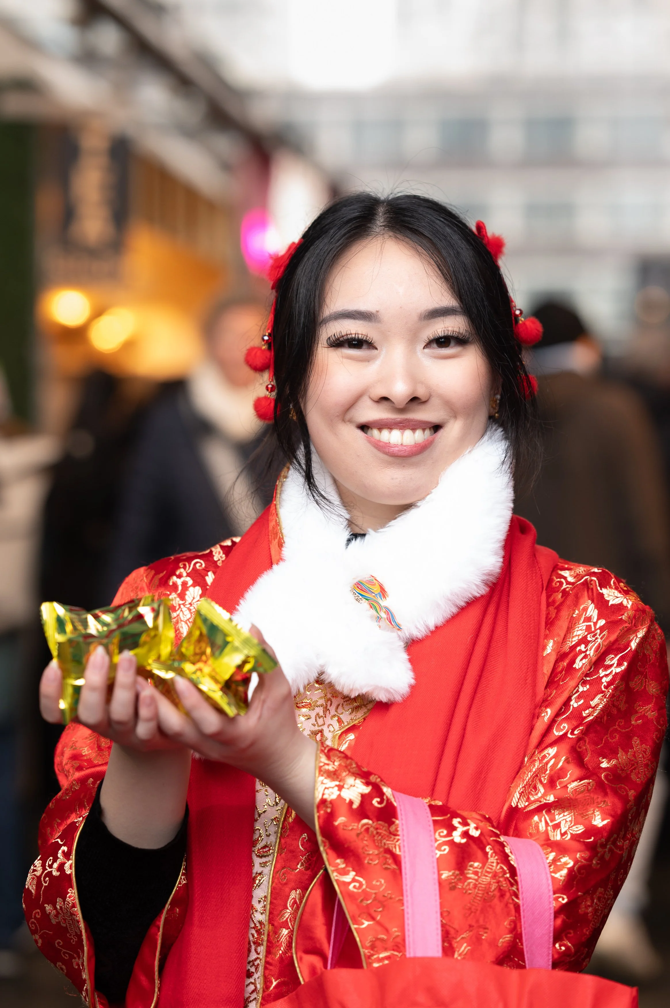 A woman dressed in a traditional red Chinese outfit with gold embroidery, holding small wrapped gifts, smiling at the camera, with a blurred indoor background.