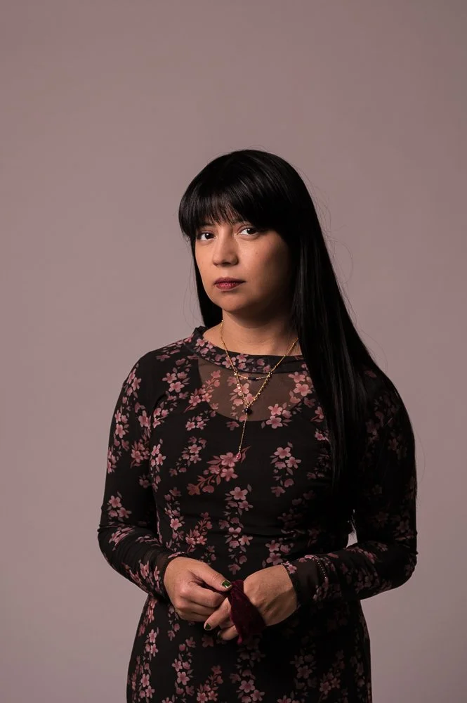 Portret van een vrouw met lang zwart haar en een zwarte jurk met roze bloemen op een eenvoudige grijze achtergrond.