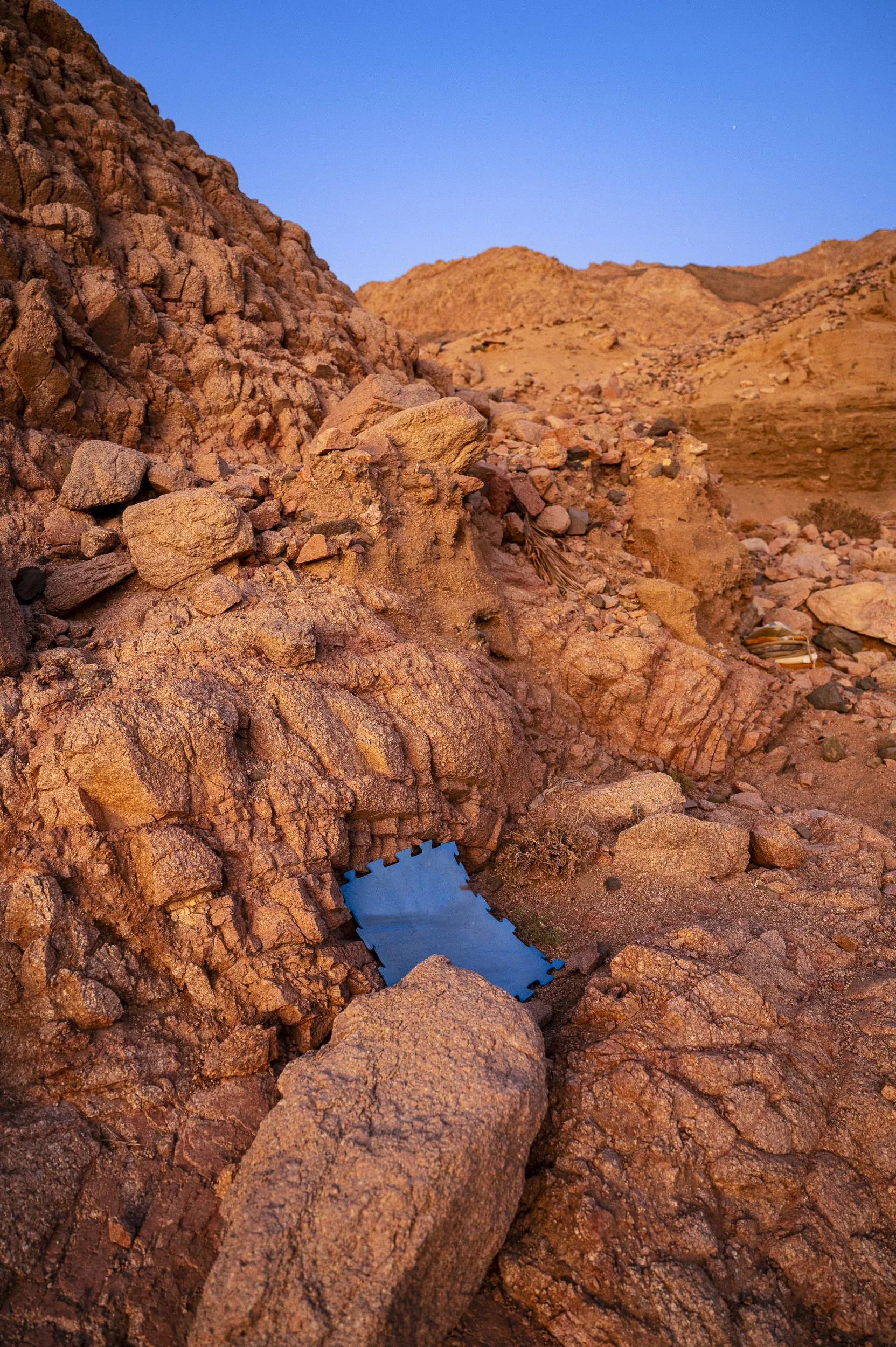 A rocky desert landscape with reddish-brown stones and a small black object near a blue square patch of tarp underneath loose rocks.