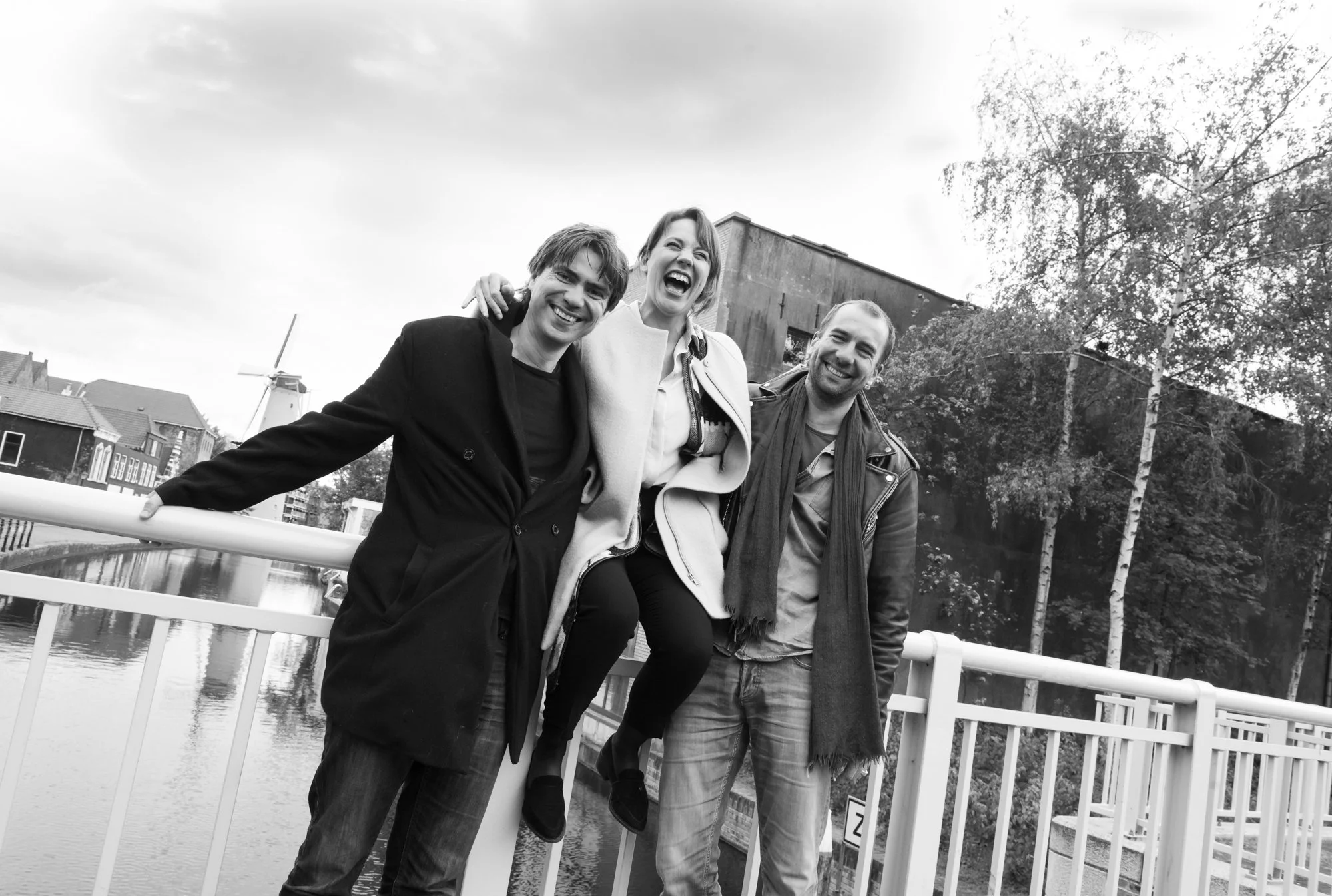 Drie mensen glimlachen en lachen terwijl ze op een brug staan, met een kanaal en huizen op de achtergrond, in een zwart-wit foto.