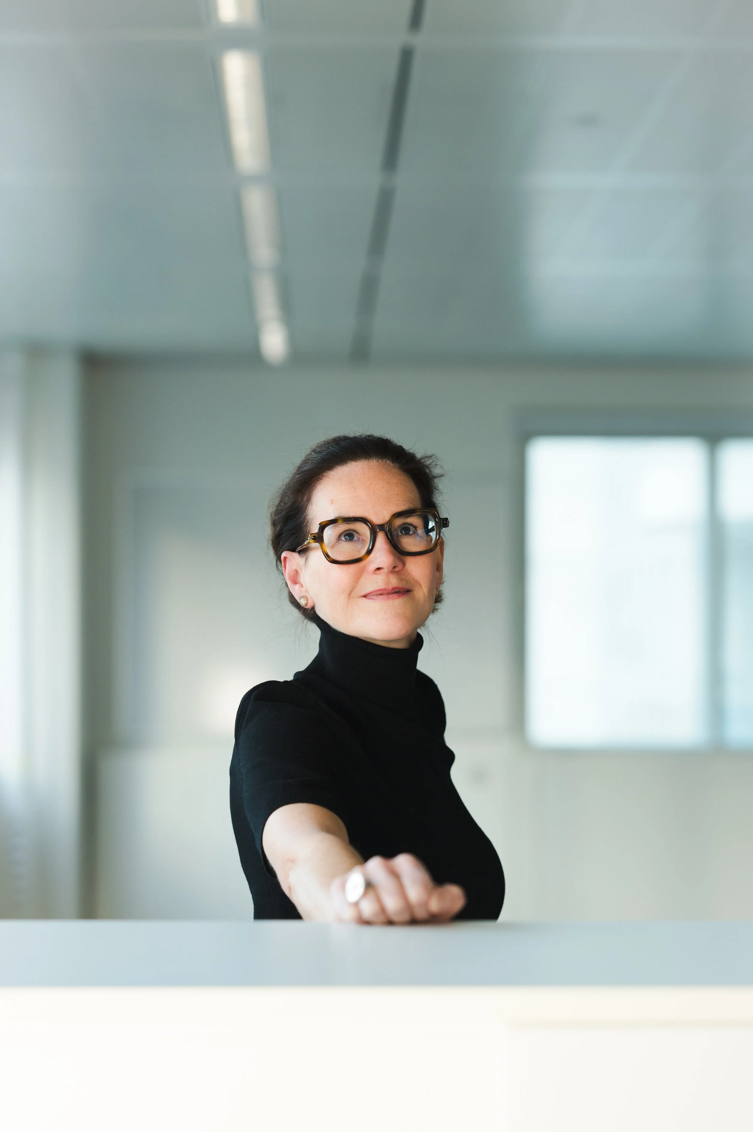 A woman with glasses in a black turtleneck extending her hand forward in an office setting.