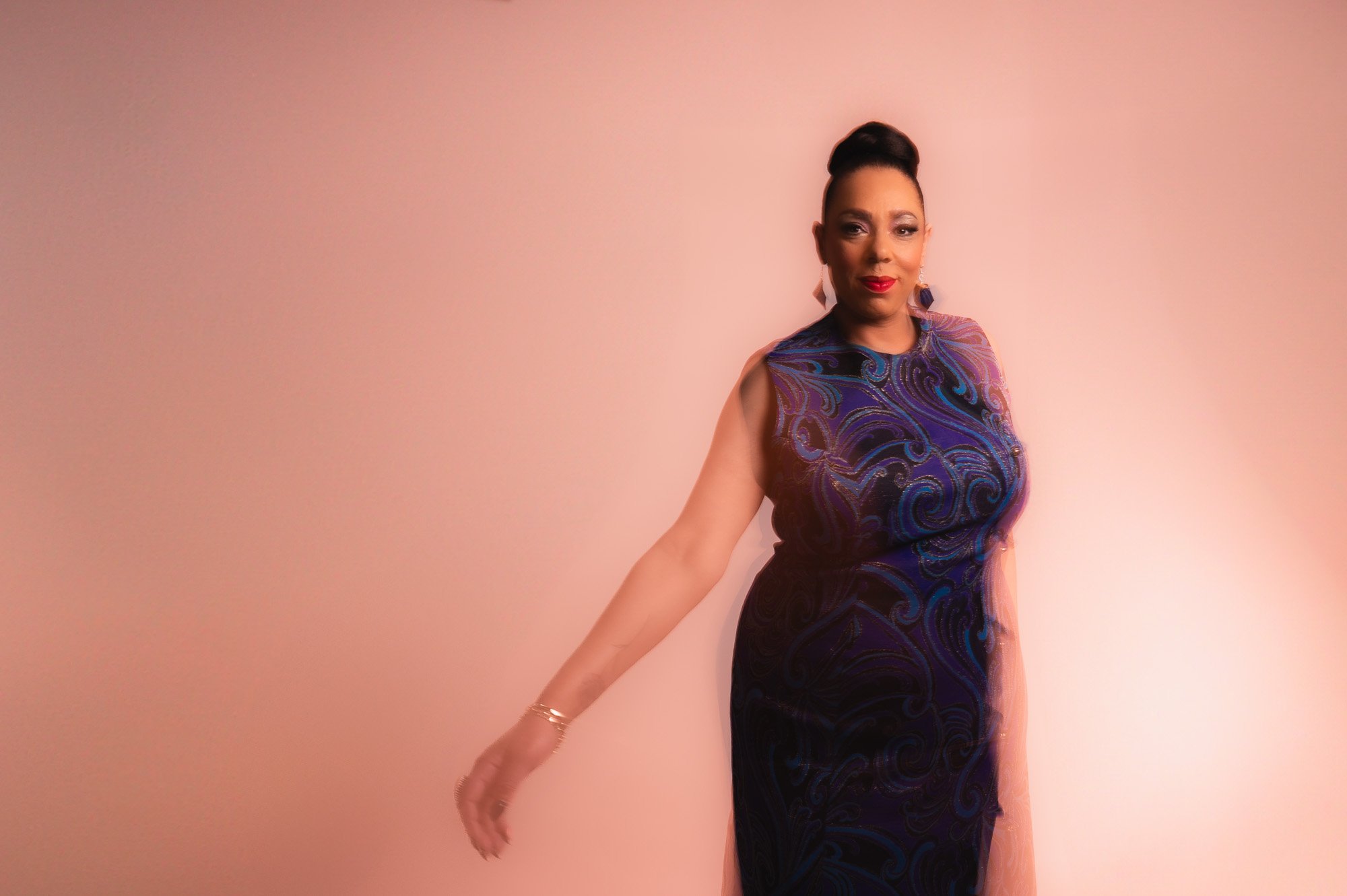 A woman in a dark blue and purple patterned evening gown, standing against a soft pink background, with her hair styled up and wearing red lipstick and earrings.