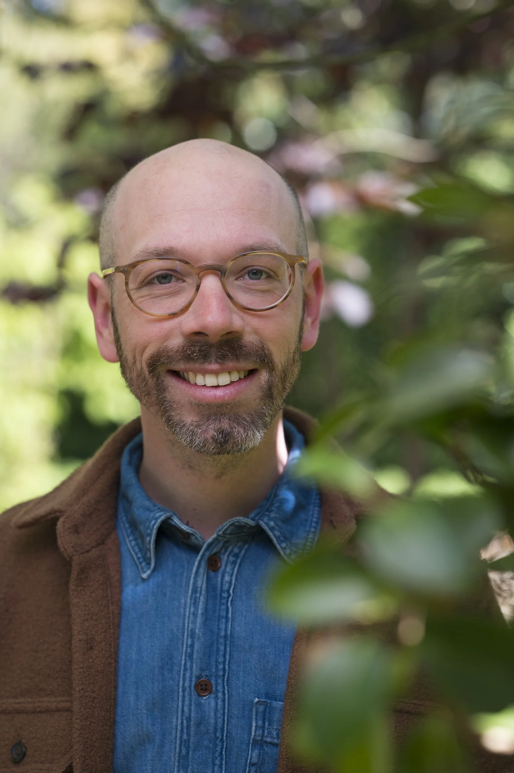 Close-up van een lachende man met brillen, blauwe overhemd en bruin jas, omringd door groene bladeren.