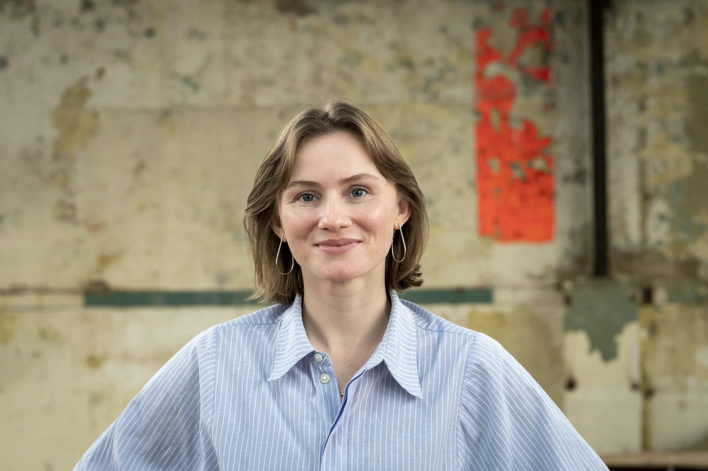 Portret van een jonge vrouw met blauwe ogen en schouderlang lichtbruin haar, die een lichte blauwe en wit gebreide blouse draagt. Ze staat voor een oude muur met verf die afbladdert, met een rode decoratieve muurhanger op de achtergrond.
