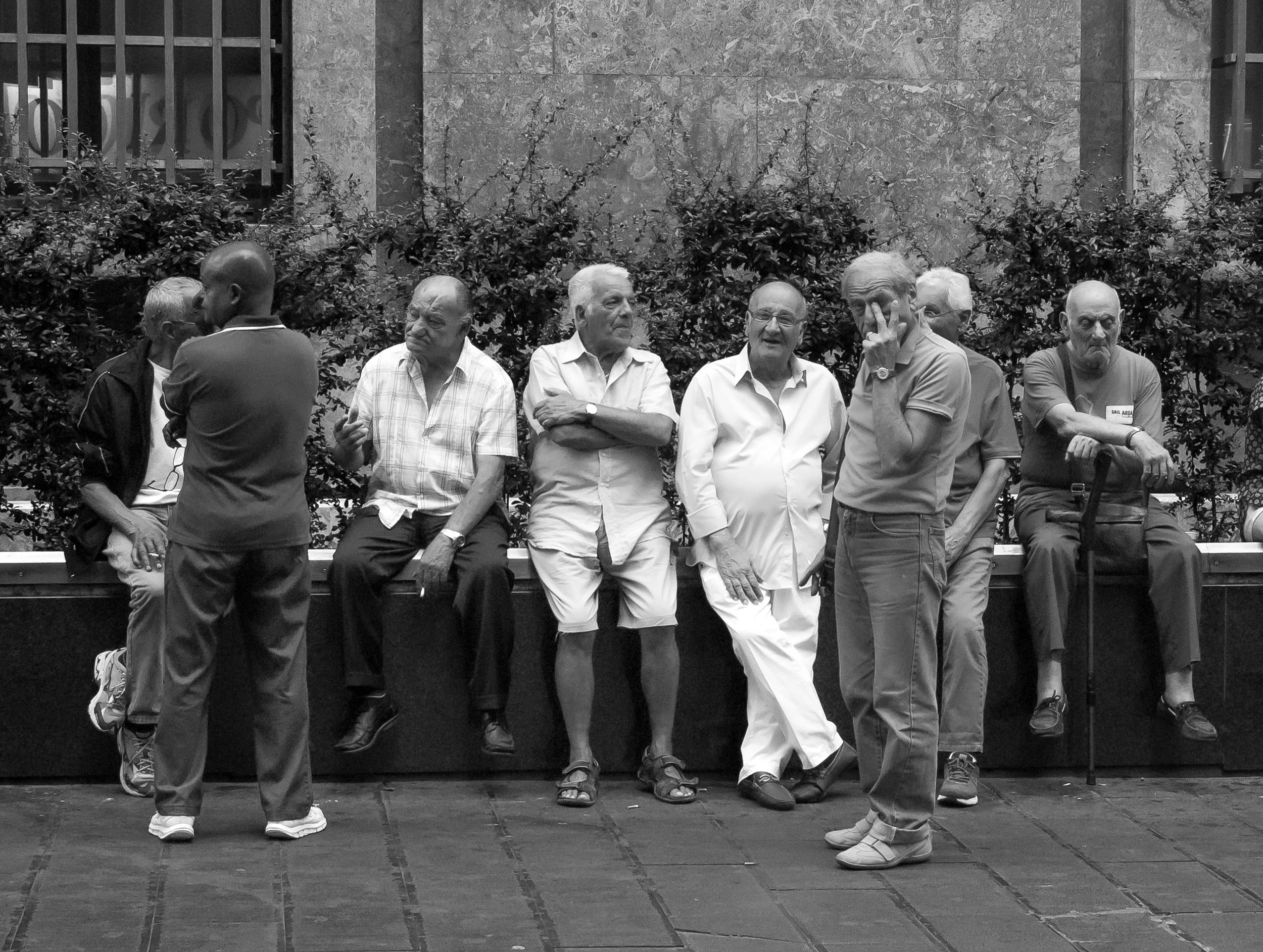Group of men sitting and standing on a bench in an outdoor urban setting, engaged in conversation and relaxed.