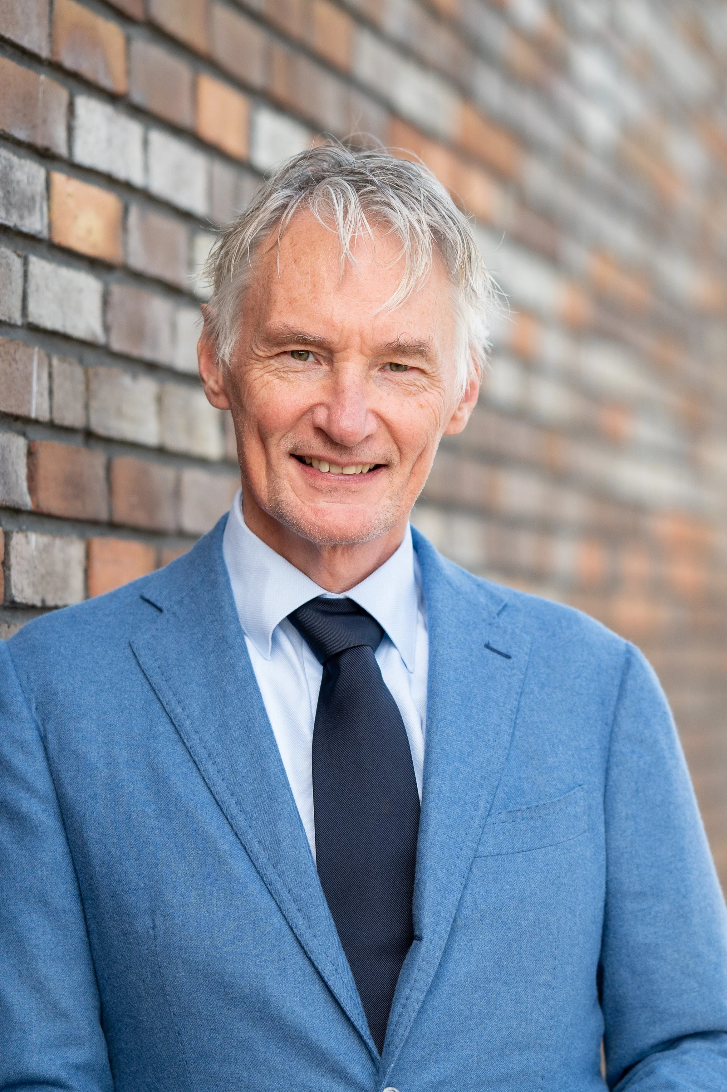 Man in een blauwe jas, witte overhemd en zwarte stropdas, lachend voor een bakstenen muur.