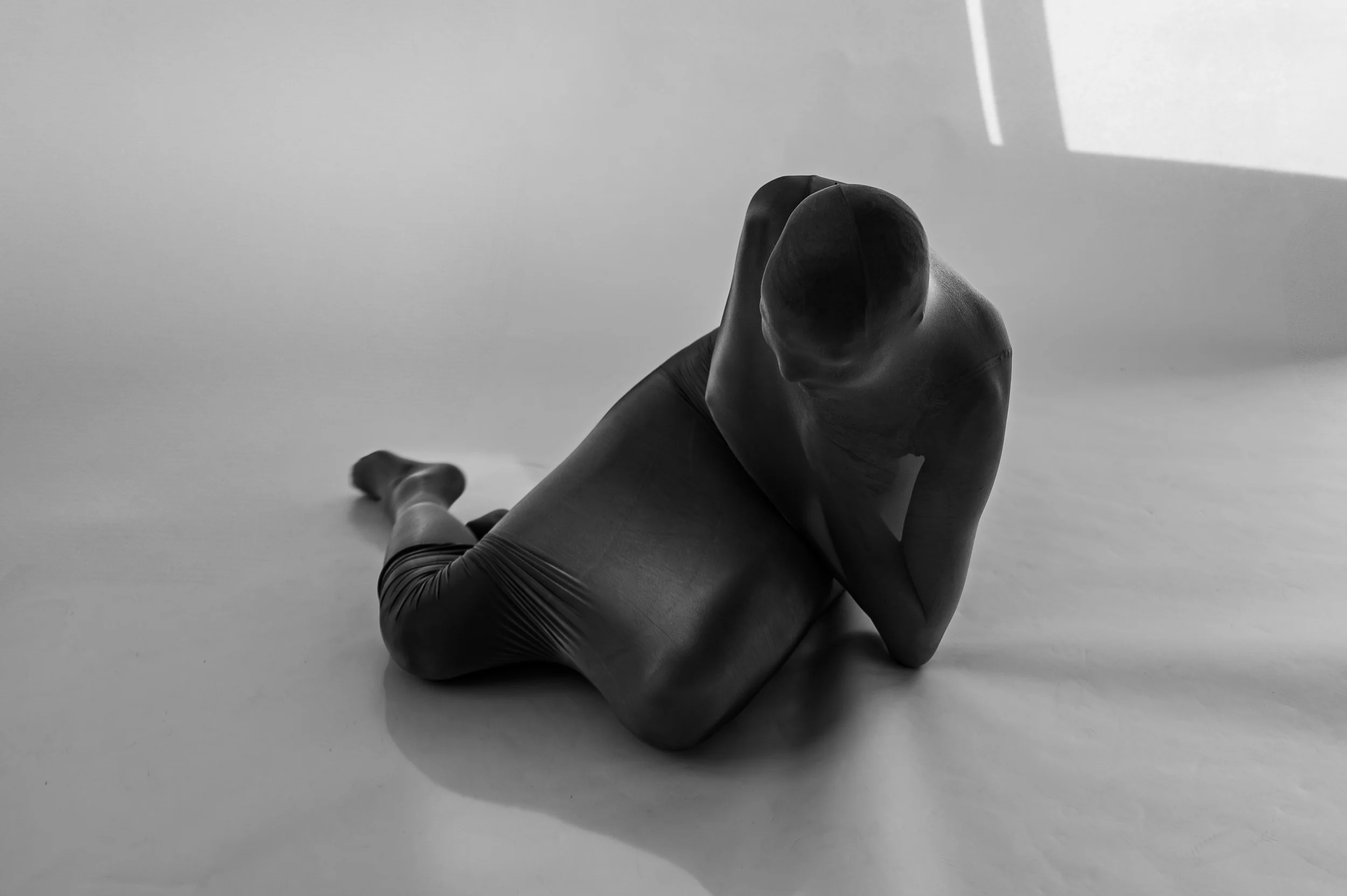 A person wearing a tight, sleeveless outfit kneeling on a smooth surface with their head bowed and arms covering their face, in a black and white photograph.