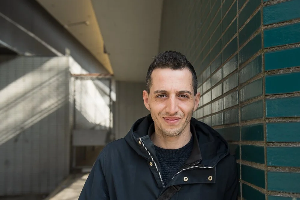 Man staat buiten naast een muur met blauwe tegels, glimlacht naar de camera.