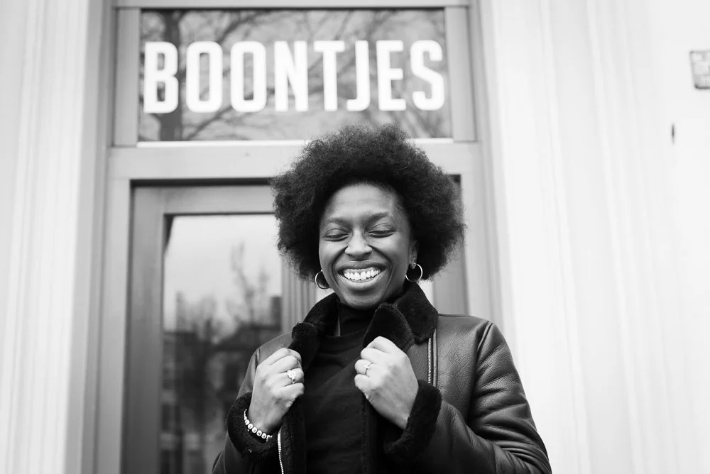A smiling woman with an afro hairstyle standing in front of a building with a sign that says 'BOONTJES'. She is wearing a black leather jacket with a fur collar and has hoop earrings.