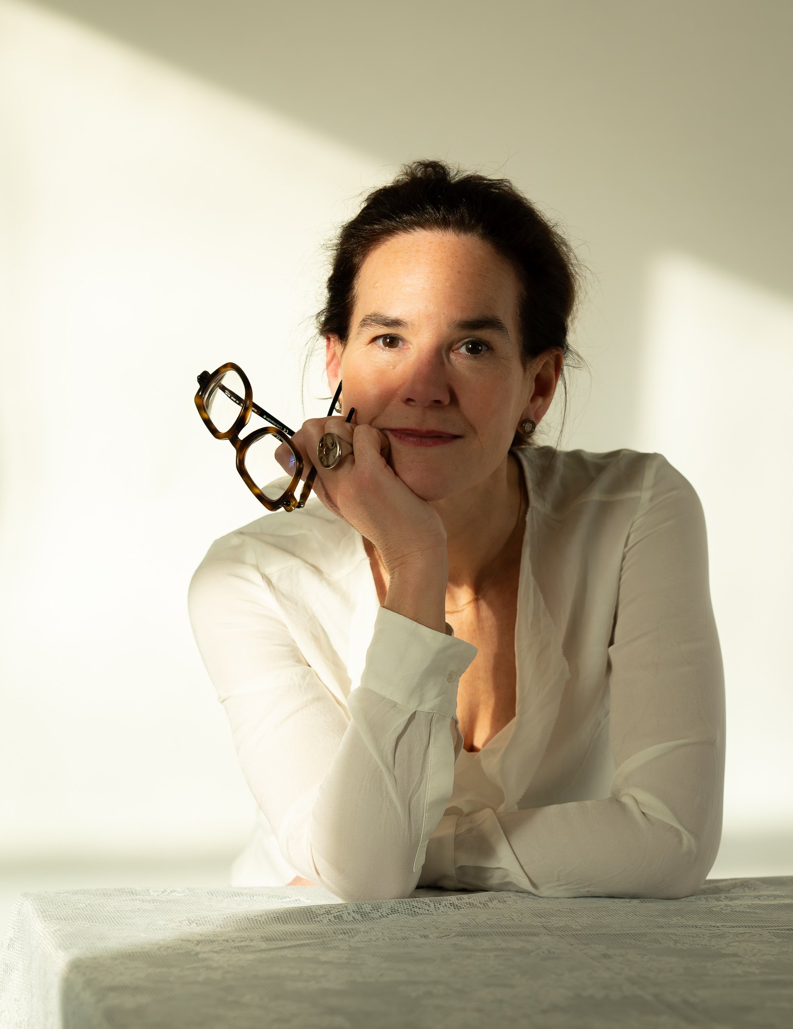 Portrait of a woman with dark hair, wearing a white blouse, holding a pair of tortoise-shell glasses to her face, sitting at a table, with a neutral background and soft lighting.