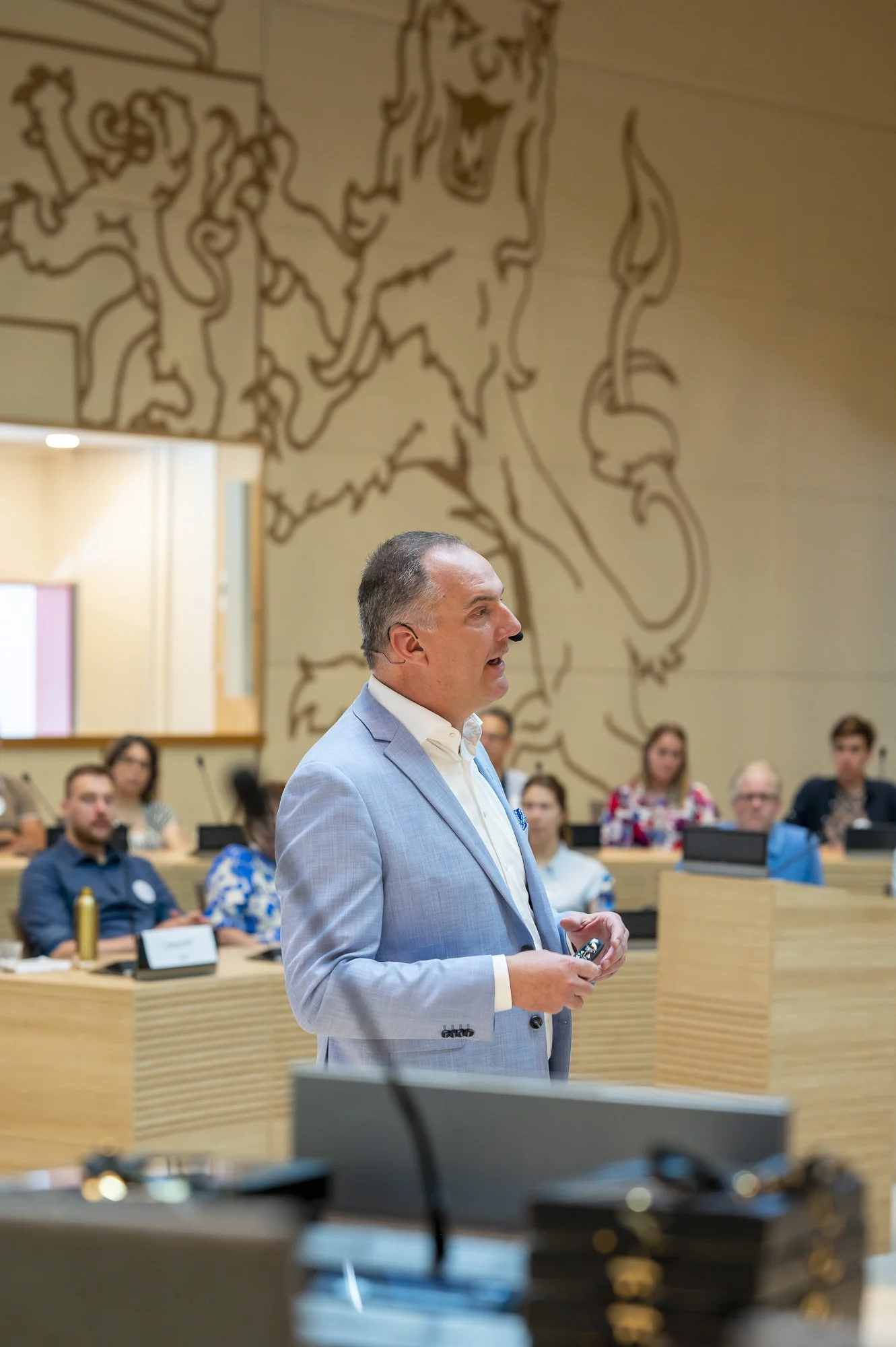 Een man in een lichtblauw pak geeft een presentatie in een conferentiezaal met een muurschildering van een leeuw op de achtergrond, terwijl een groep mensen hem hoort.