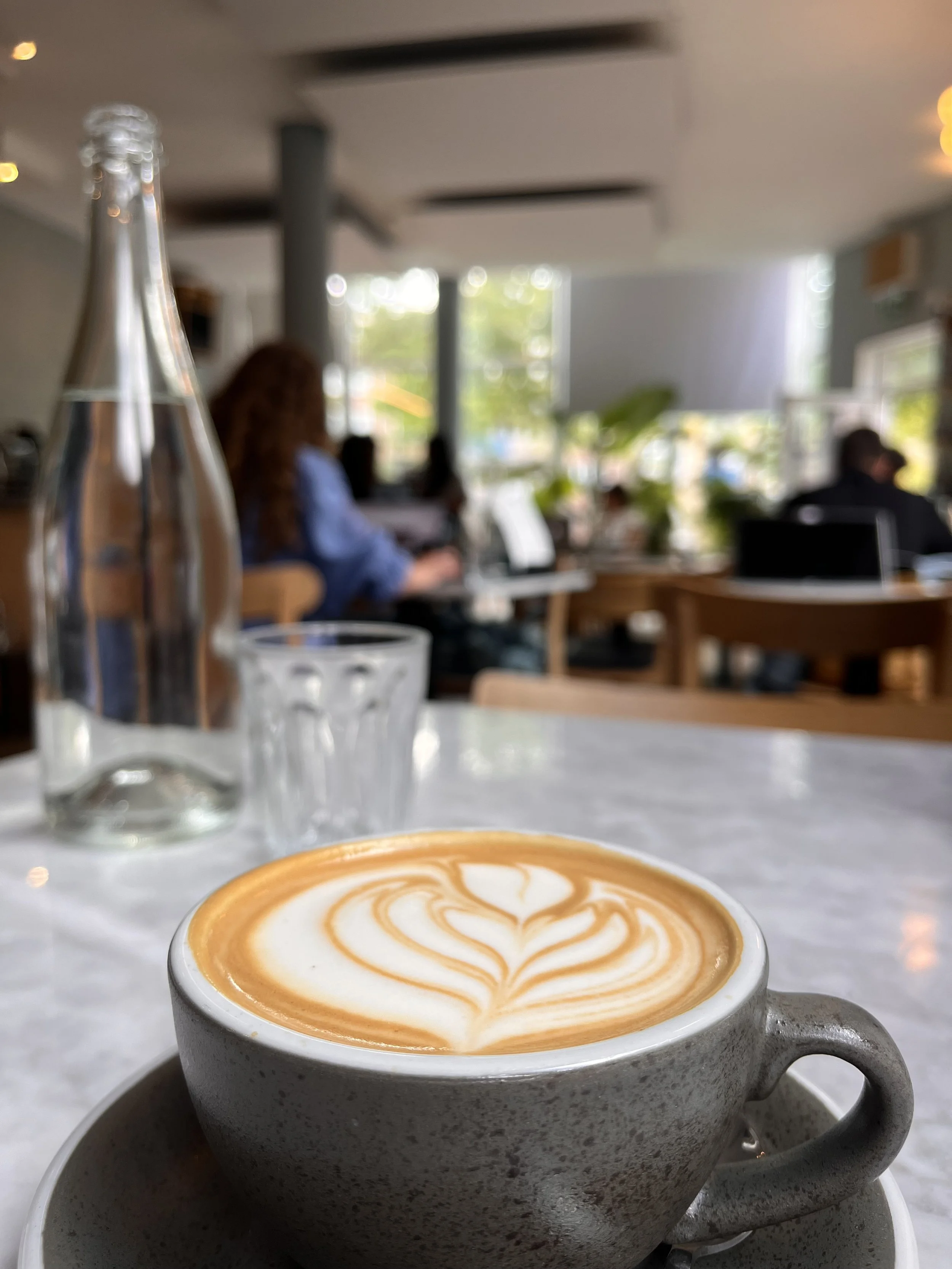 Een kopje koffie met latte art, op de voorgrond. Op de achtergrond zien we mensen die binnen in een café of restaurant zitten, met grote ramen en veel natuurlijk licht. FOTOGRAFIE.