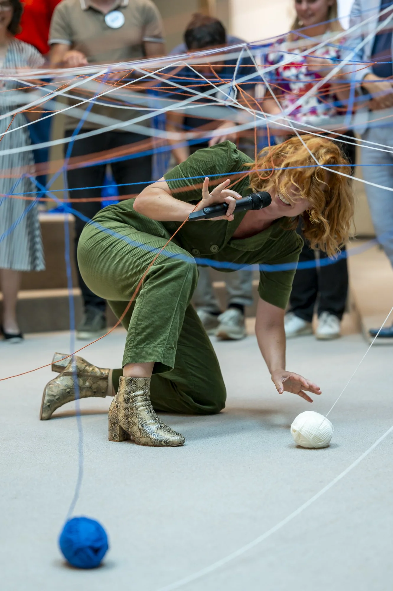 Een vrouw met glanzende laarzen en groene kleding knielt op de vloer terwijl ze een draad of touw vasthoudt. Ze glimlacht en houdt een microfoon vast in haar rechterhand. Op de vloer voor haar ligt een witte bal en er zijn meerdere gekleurde draadkaa