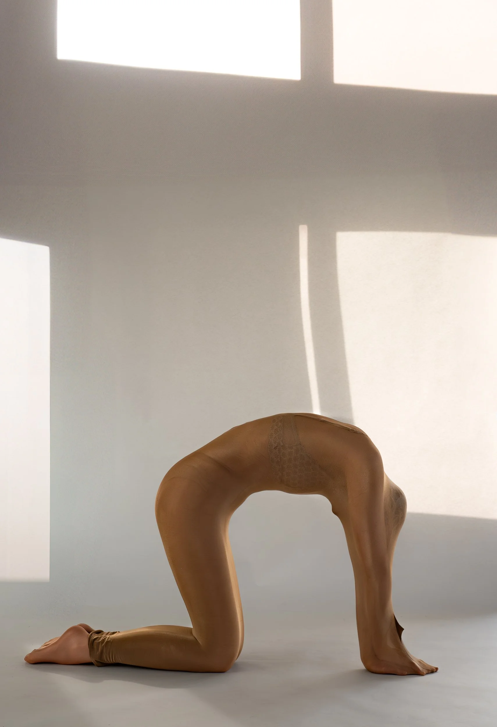 A person in tan tights kneeling with their head and arms on the floor, creating an arch with their body, against a white background with shadow patterns from window light.