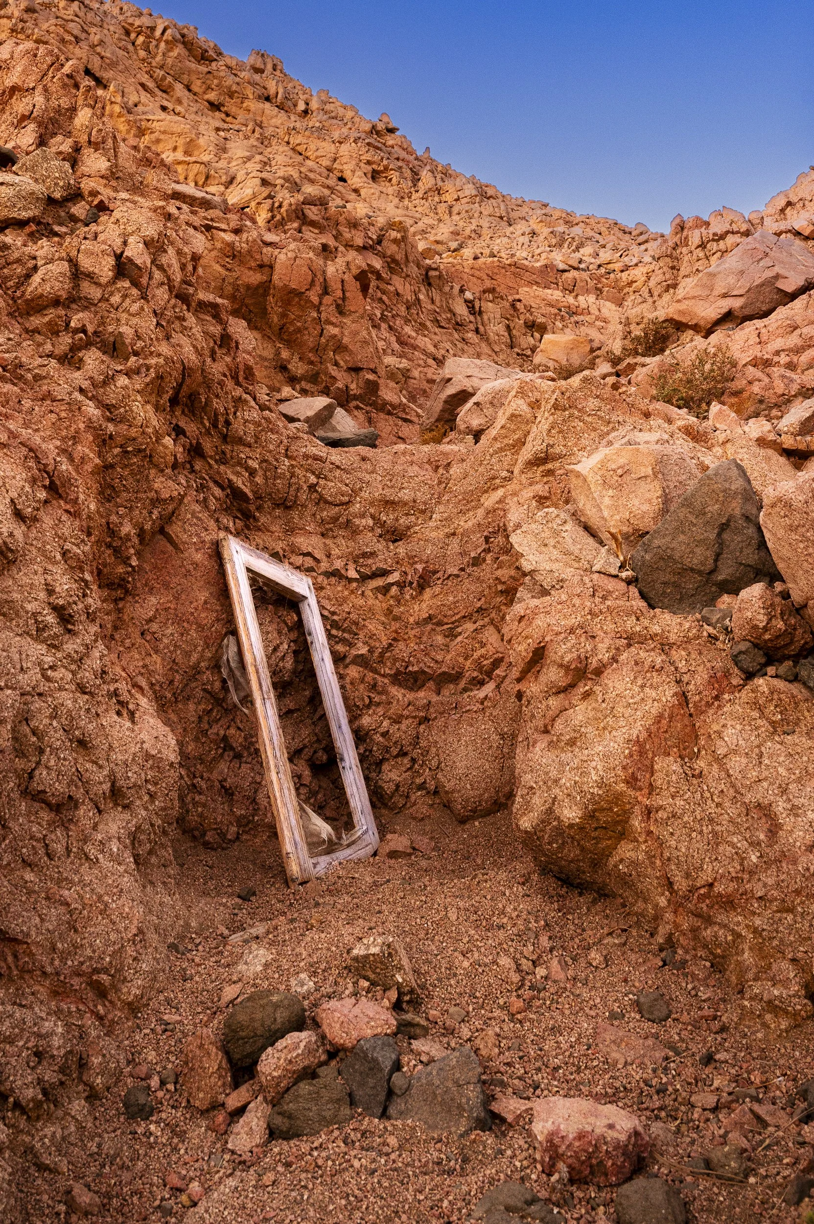 Desert landscape with reddish-brown rocky cliffs and a discarded weathered wooden picture frame leaning against the rocks.