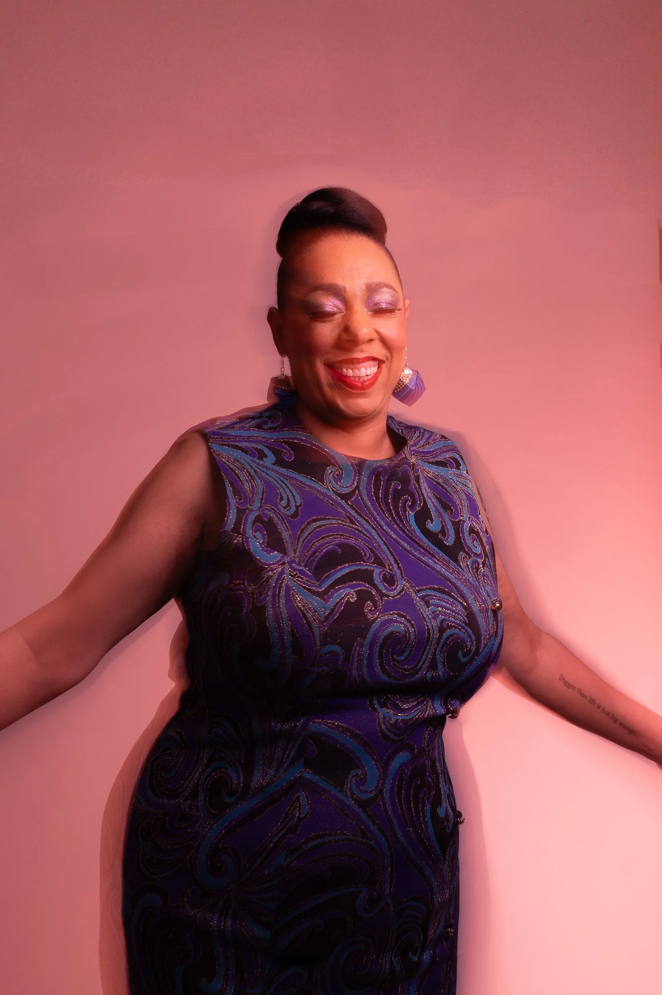 A woman smiling, wearing a sleeveless purple dress with intricate swirling patterns, standing against a pink background.