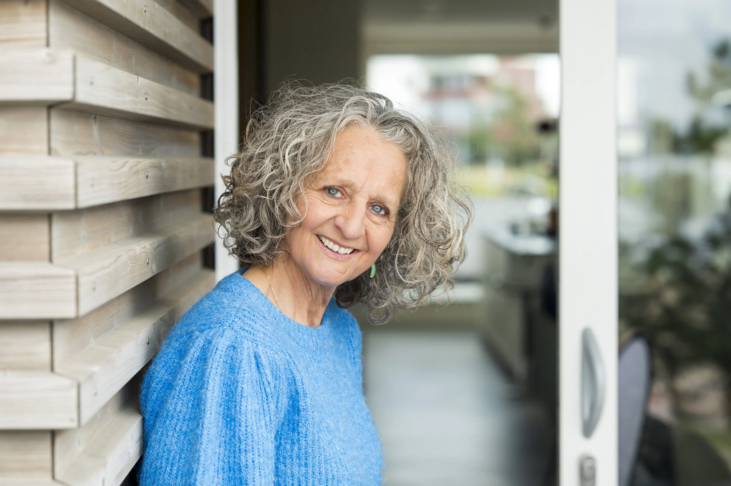 Lachende oudere vrouw met grijs, gekruld haar, die een blauwe gebreide trui draagt, staat buiten, leunt tegen een houten muur en kijkt naar de camera.