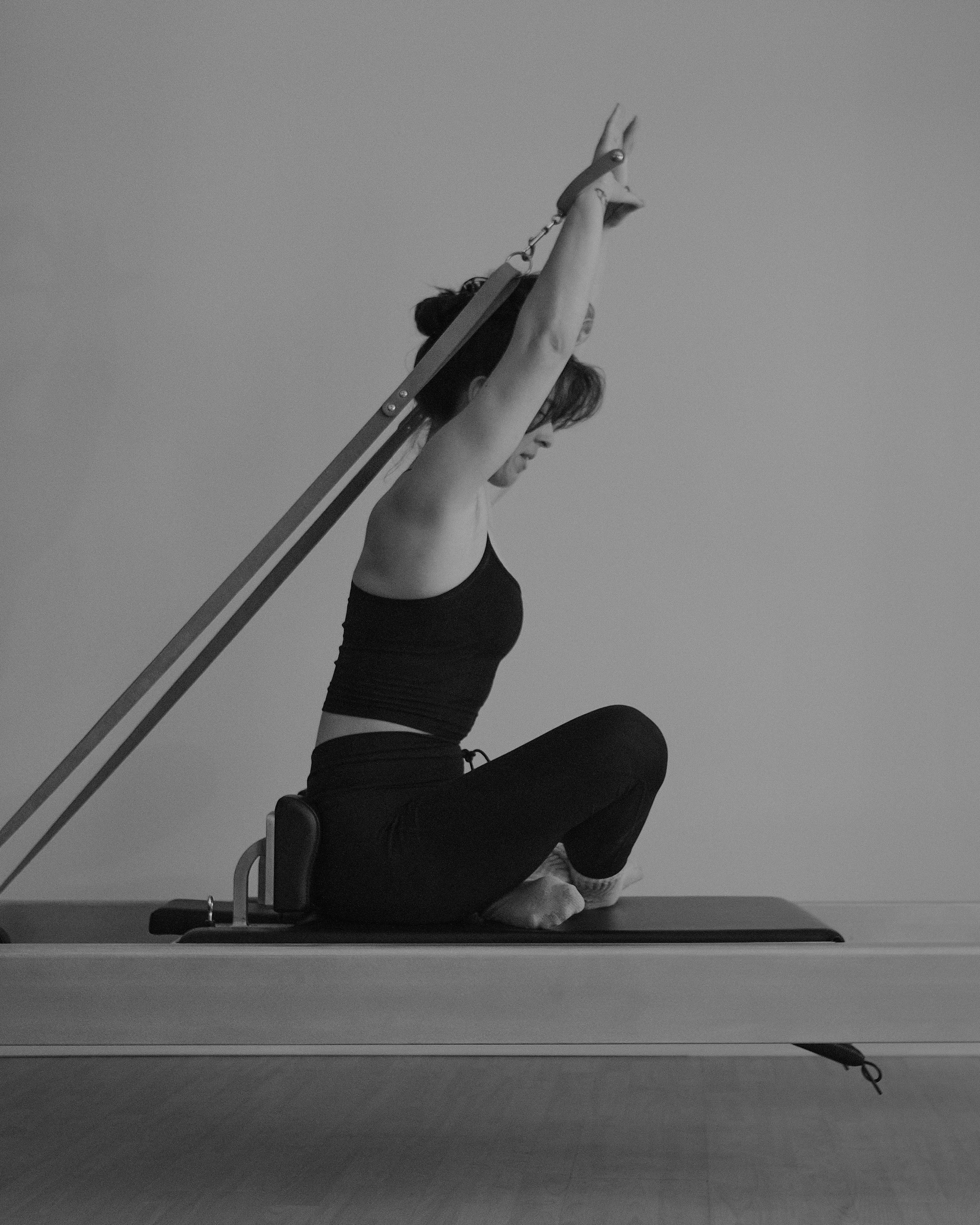 A person kneeling on a yoga mat, leaning back and reaching up with both arms, with a support frame behind them, against a plain background.