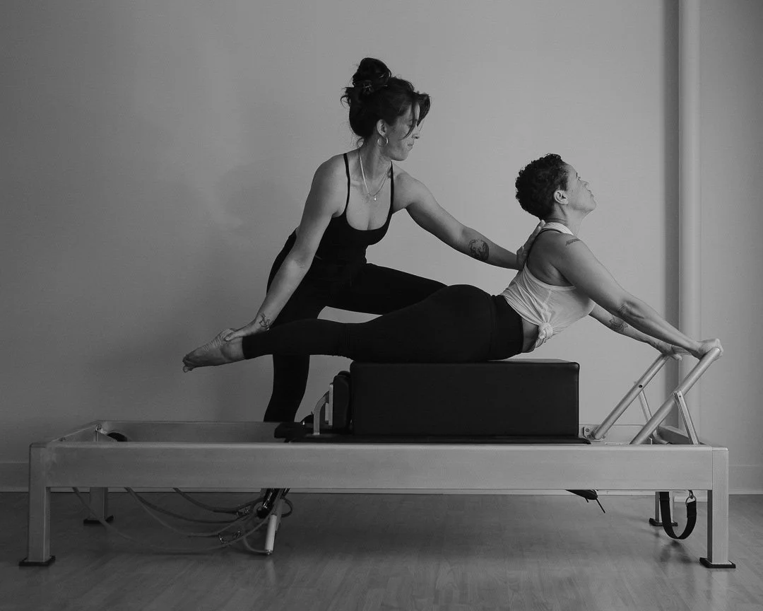 Two women practicing pilates on a reformer machine, one assisting the other with an extended stretch.