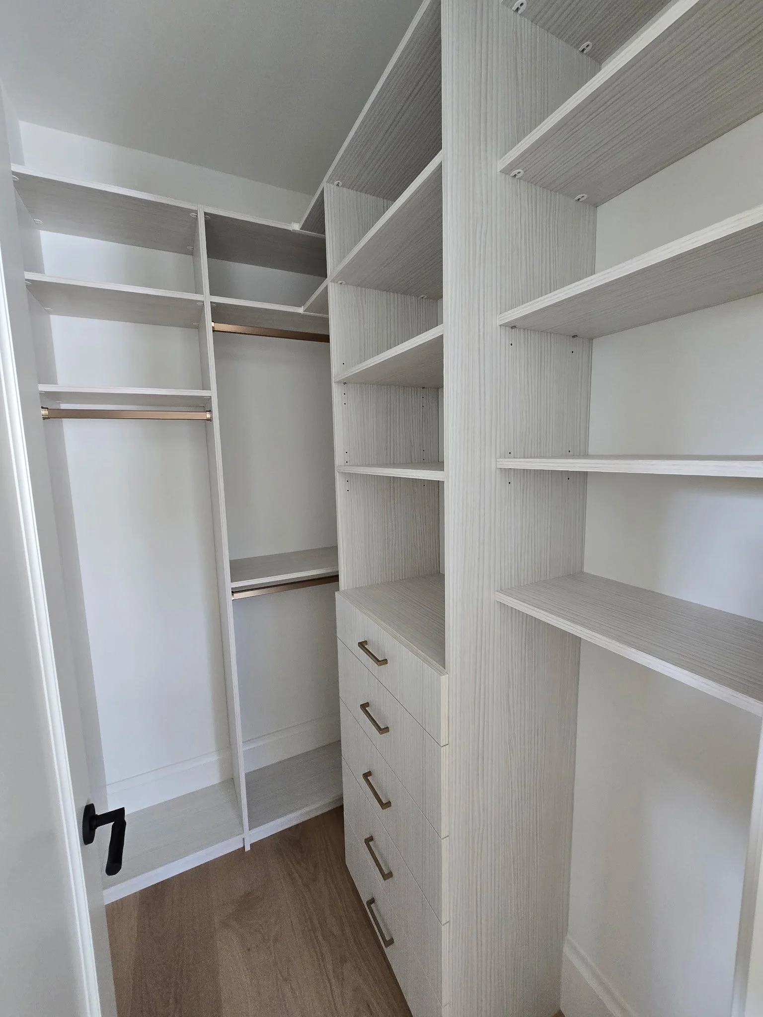 Empty walk-in closet with white textured shelving units, drawers, golden handles rods, and wooden flooring.