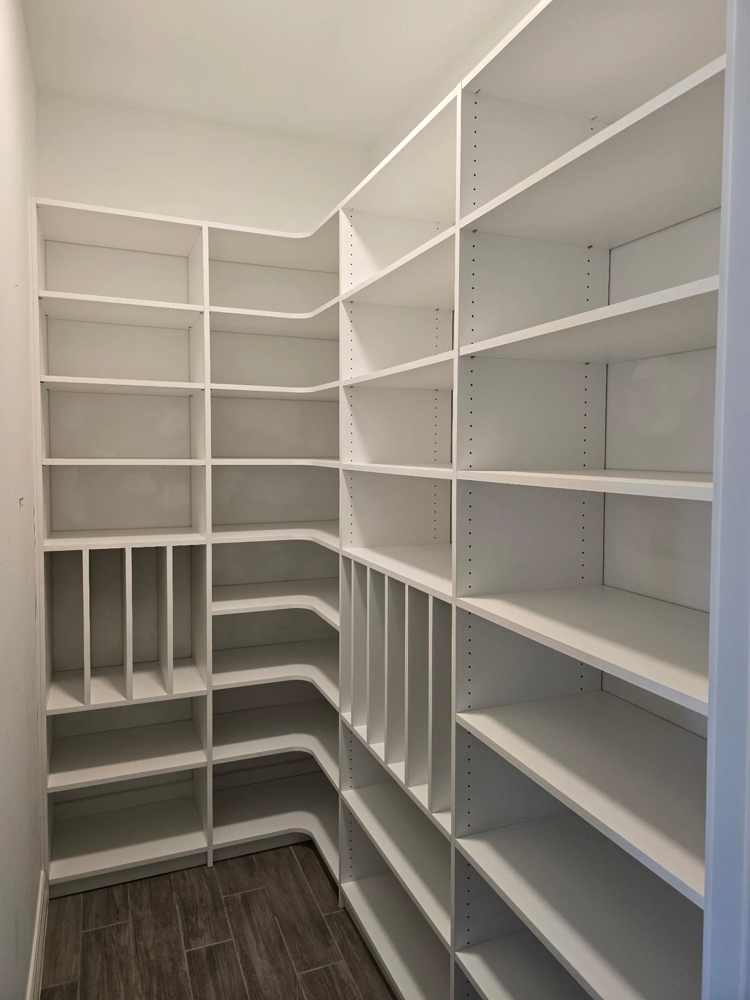 Empty white built-in corner pantry with multiple shelves and compartments, located in a room with dark wood flooring.