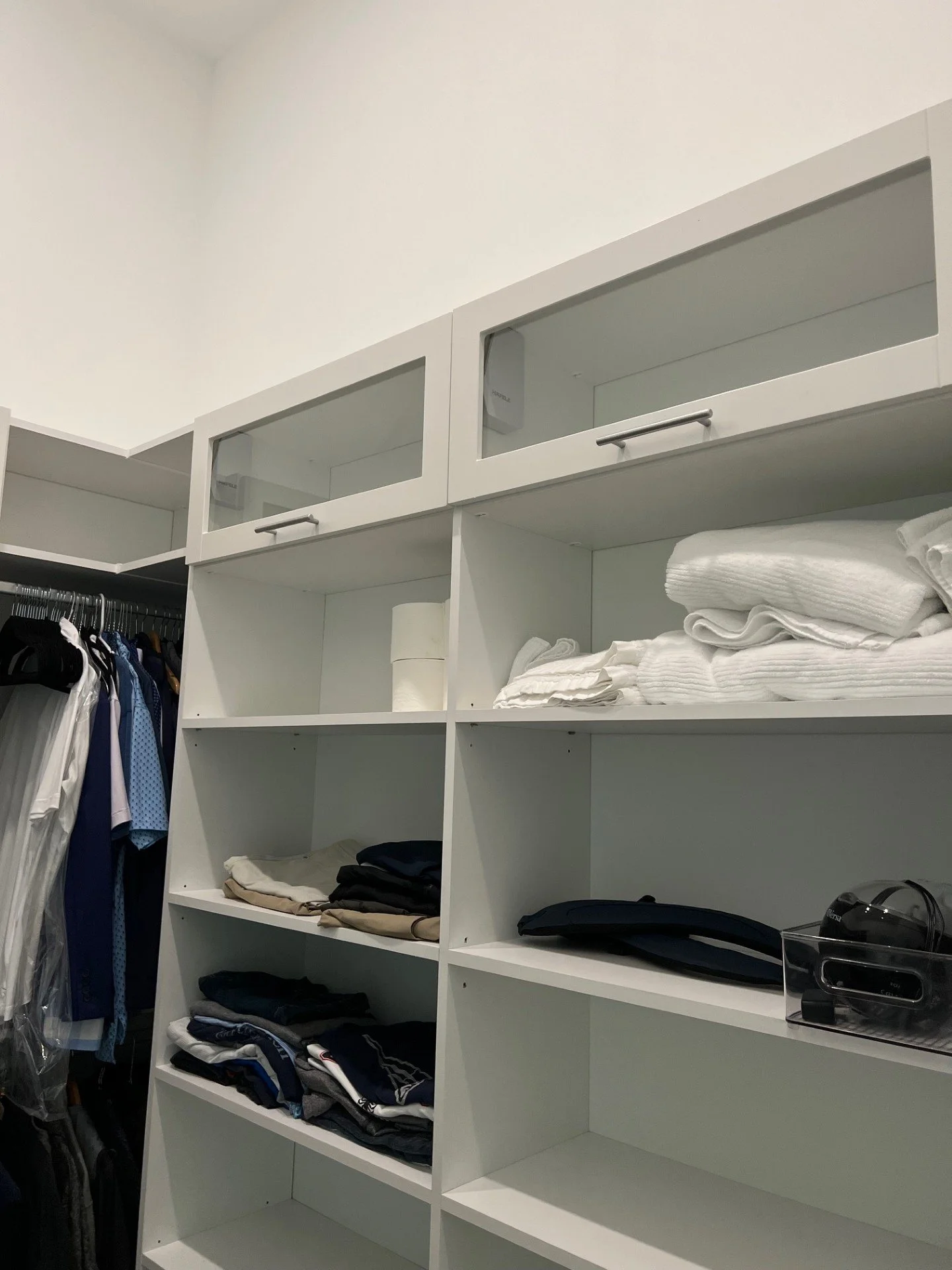 Empty white shelving unit with some folded clothes and a box on the shelves, next to hanging clothes.