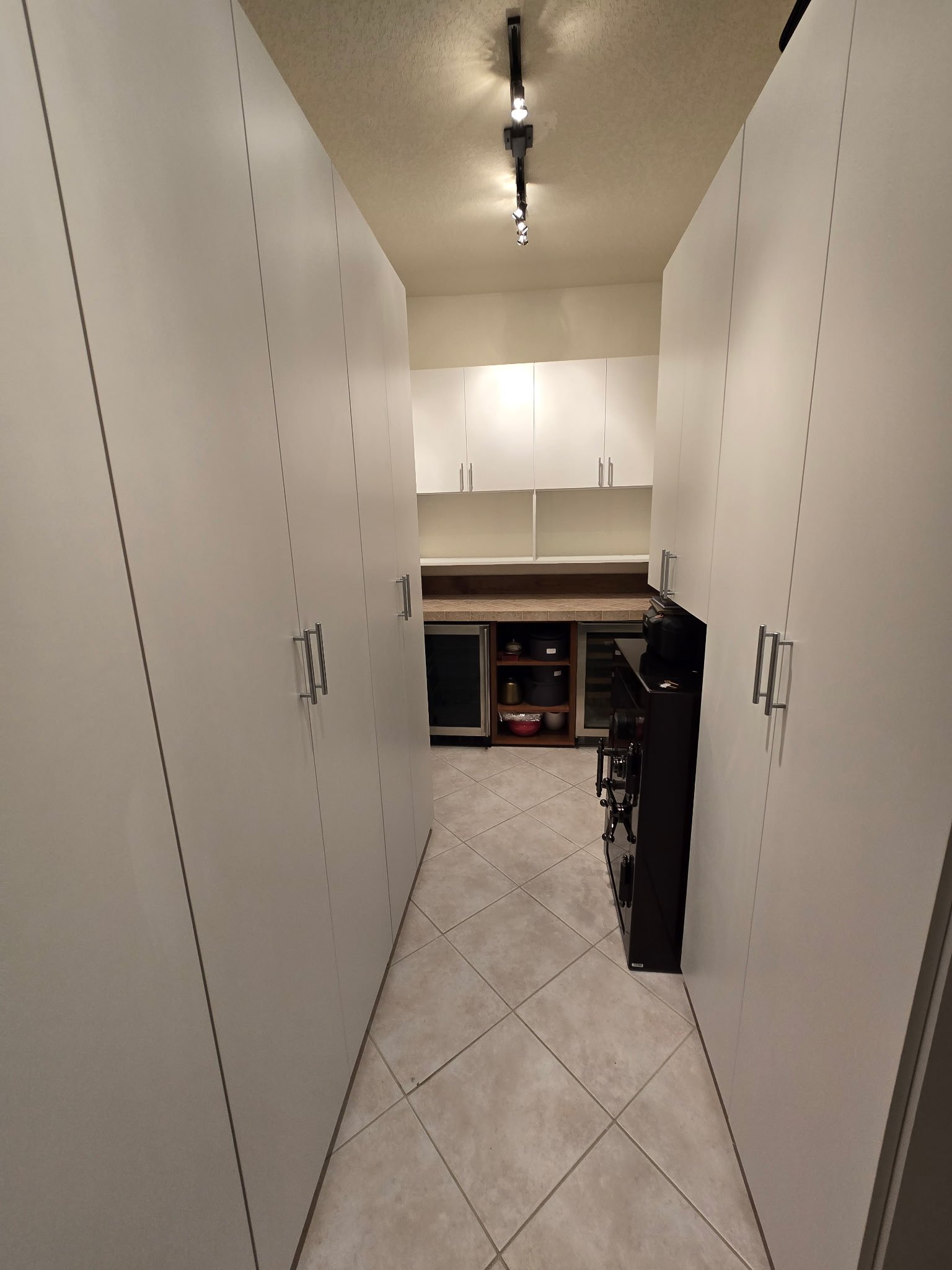 A narrow kitchen with beige tiled floor, white cabinets on both sides, and a dark countertop at the end with glass-front mini fridge, shelves with dishes, and a small black microwave.