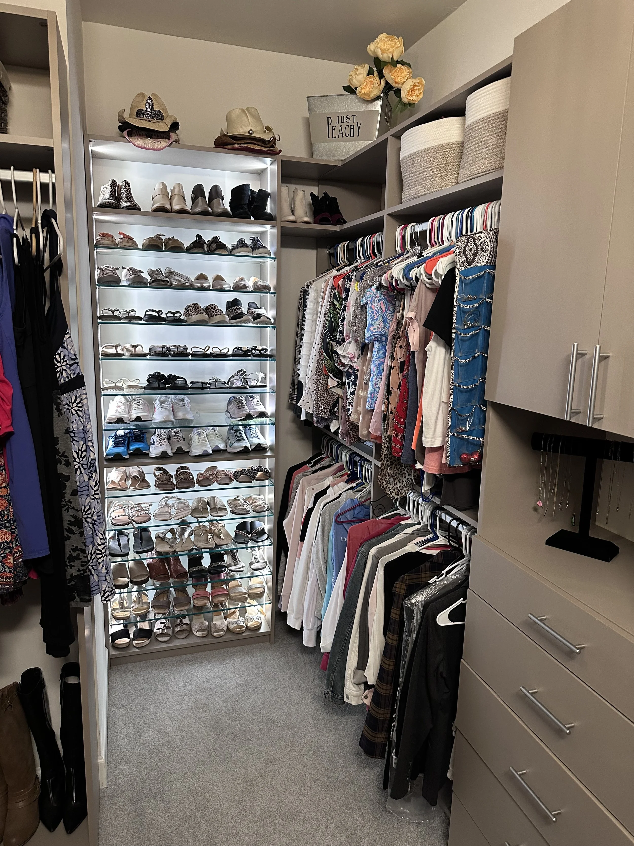 A walk-in closet with shoes on a built-in shelf, clothing on hangers, hats on top shelves, and decorative boxes and flowers on upper shelves.