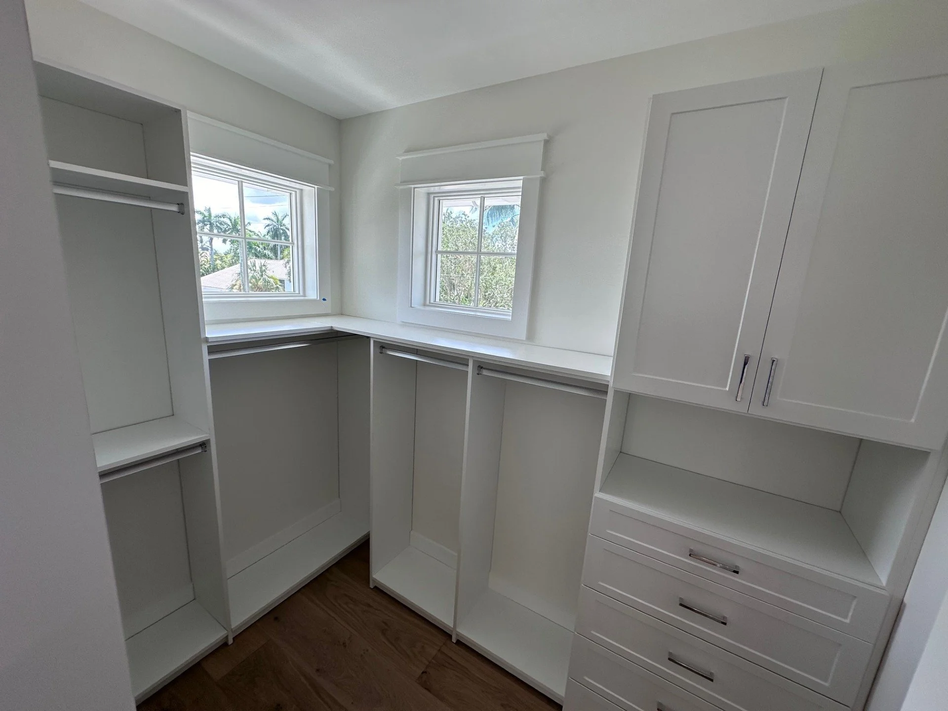 Empty white walk-in closet with built-in shelves and drawers, two windows with white trim, and hardwood flooring.
