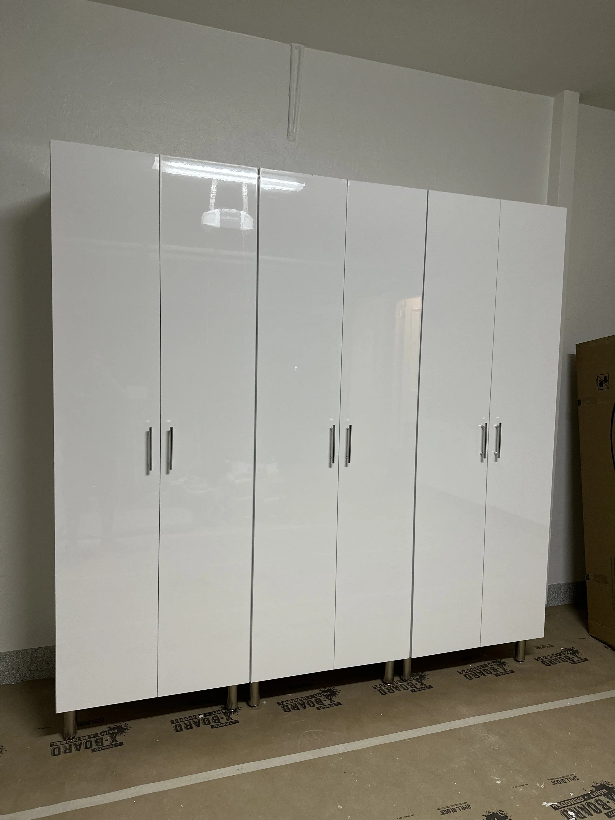 A white, glossy, multi-door storage cabinet with silver handles, standing on metal legs in a room with a concrete floor and a white painted wall.