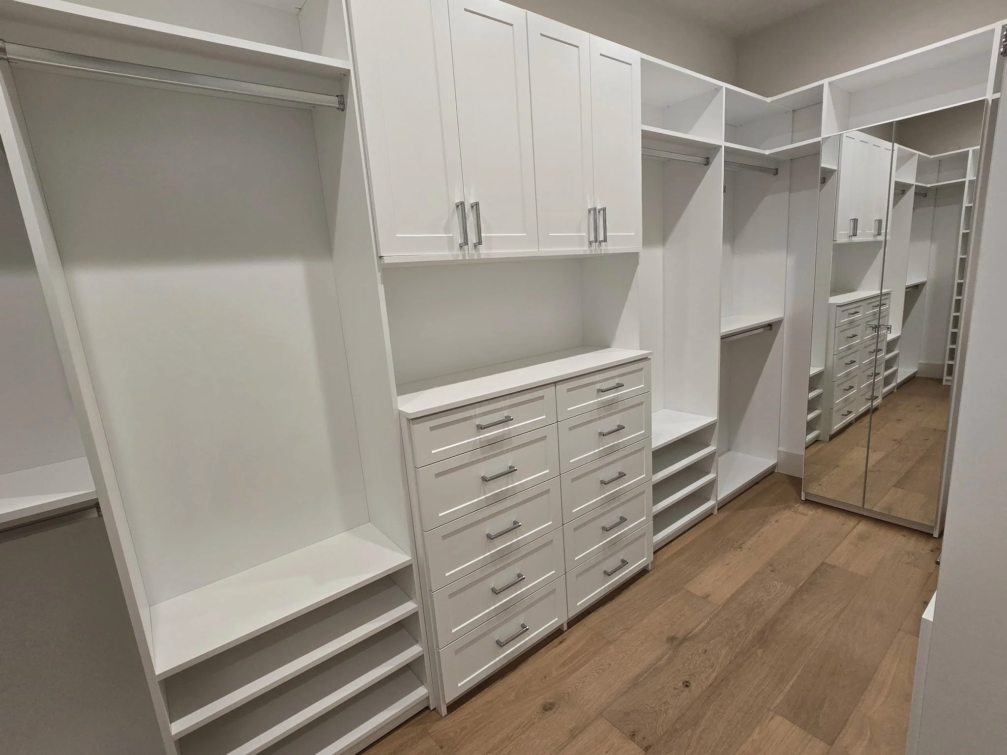 Walk-in closet with white built-in shelves, drawers, and mirrored closet doors, and hardwood flooring.