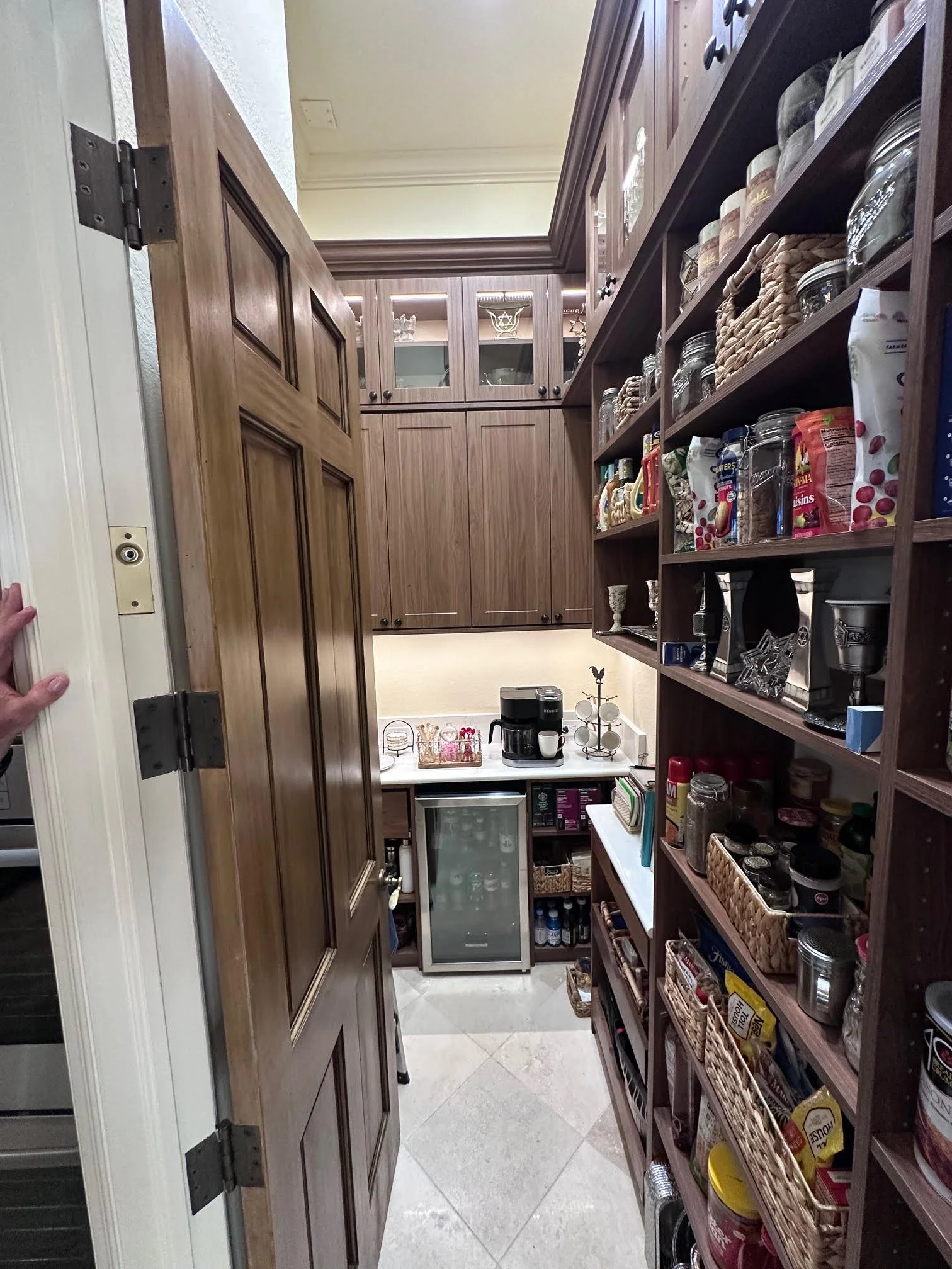 A small pantry with wooden shelves stocked with jars, cans, and baskets, a mini fridge, and a coffee station with a coffee maker and cups.