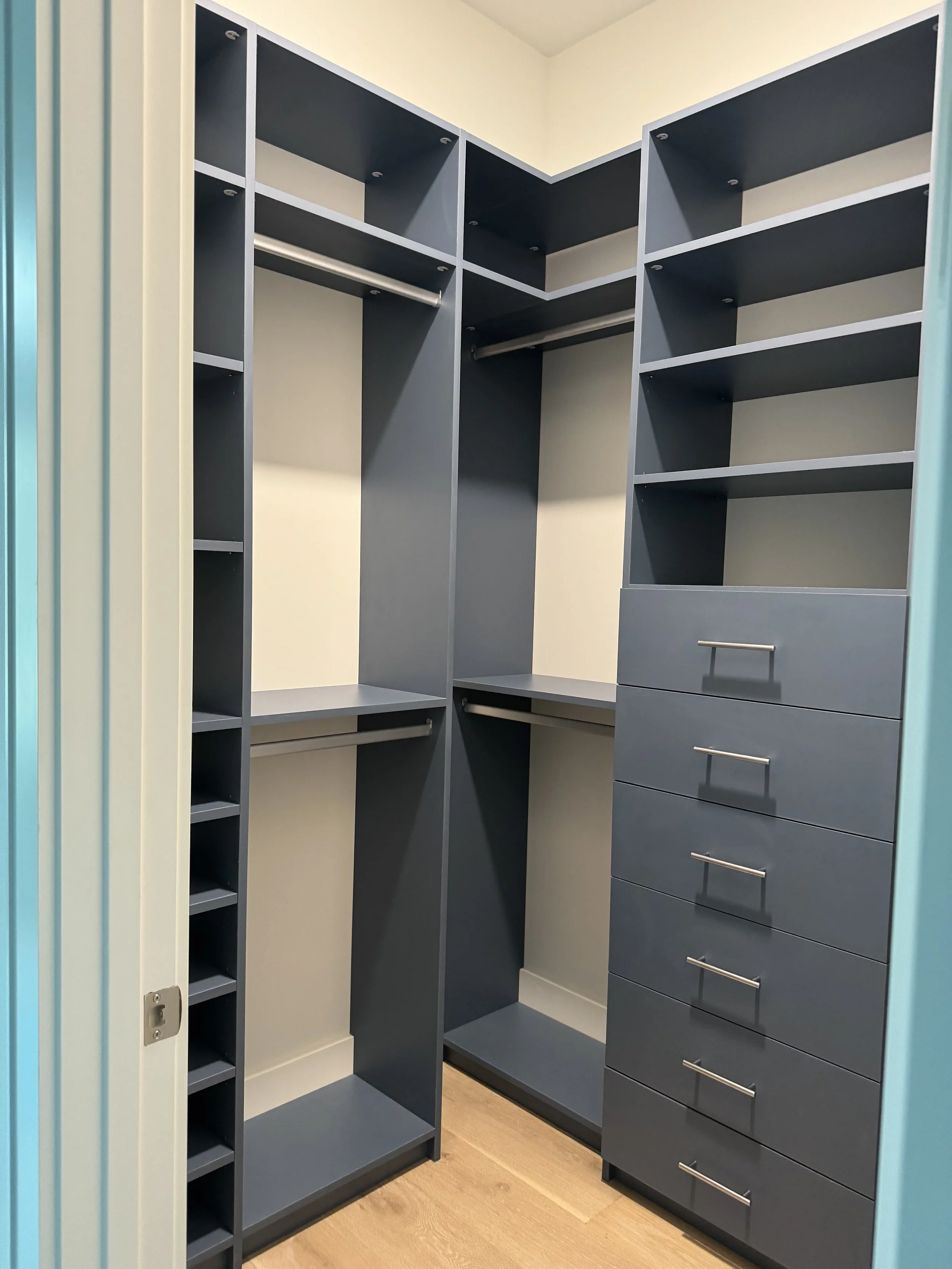 Empty walk-in closet with dark gray shelves, drawers, and hanging rods, beige walls, and light wood flooring.