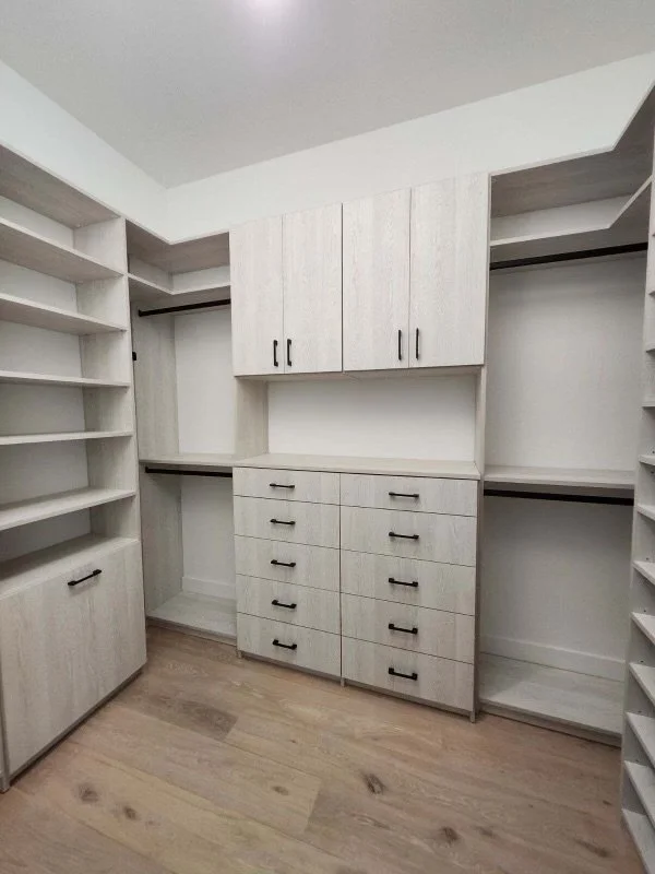 Empty walk-in closet with white built-in shelving, drawers, and hanging space, and a light wood floor.