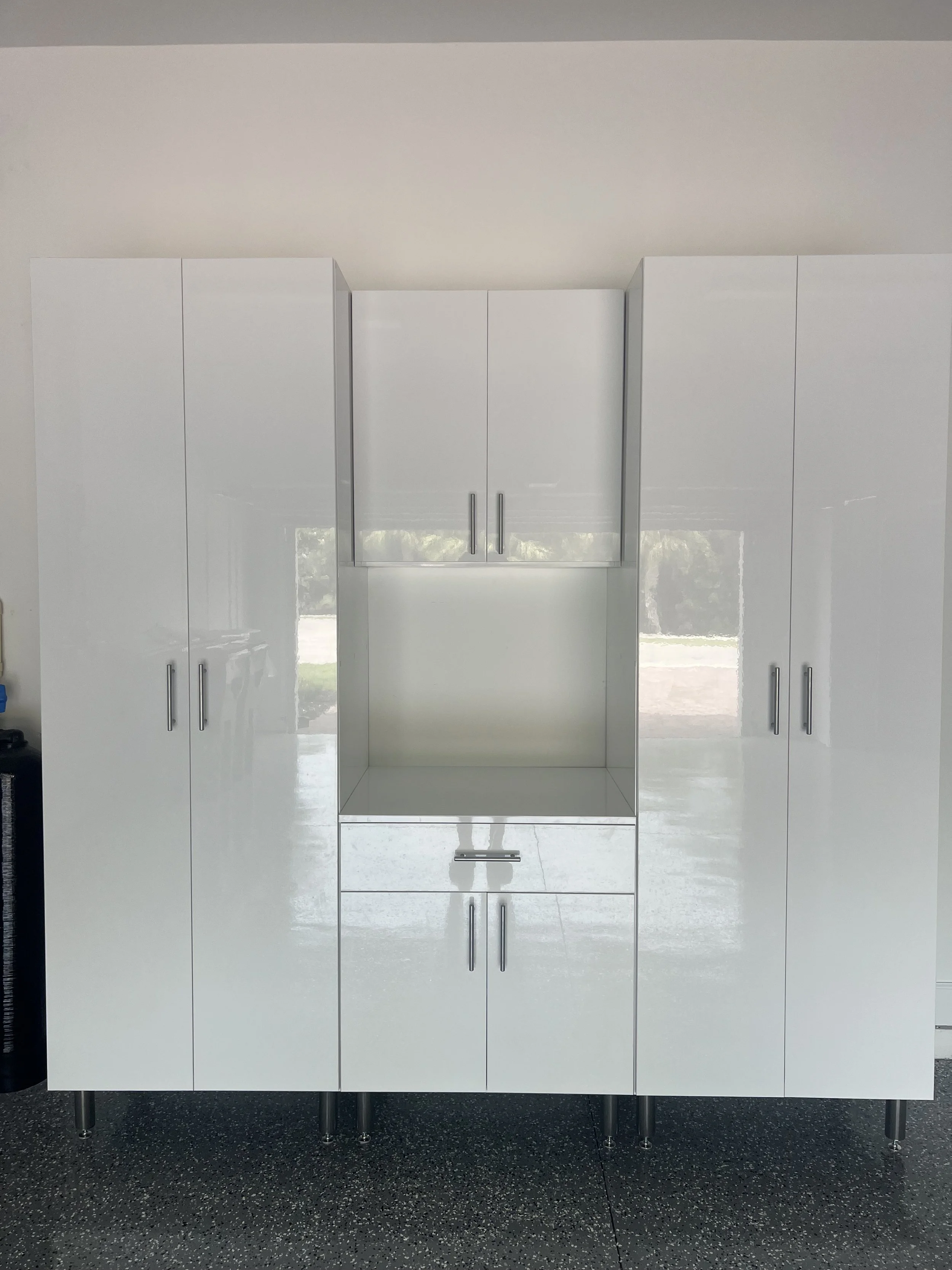 White kitchen cabinets with silver handles, designed with a central open space and drawers below, set on a black speckled floor.