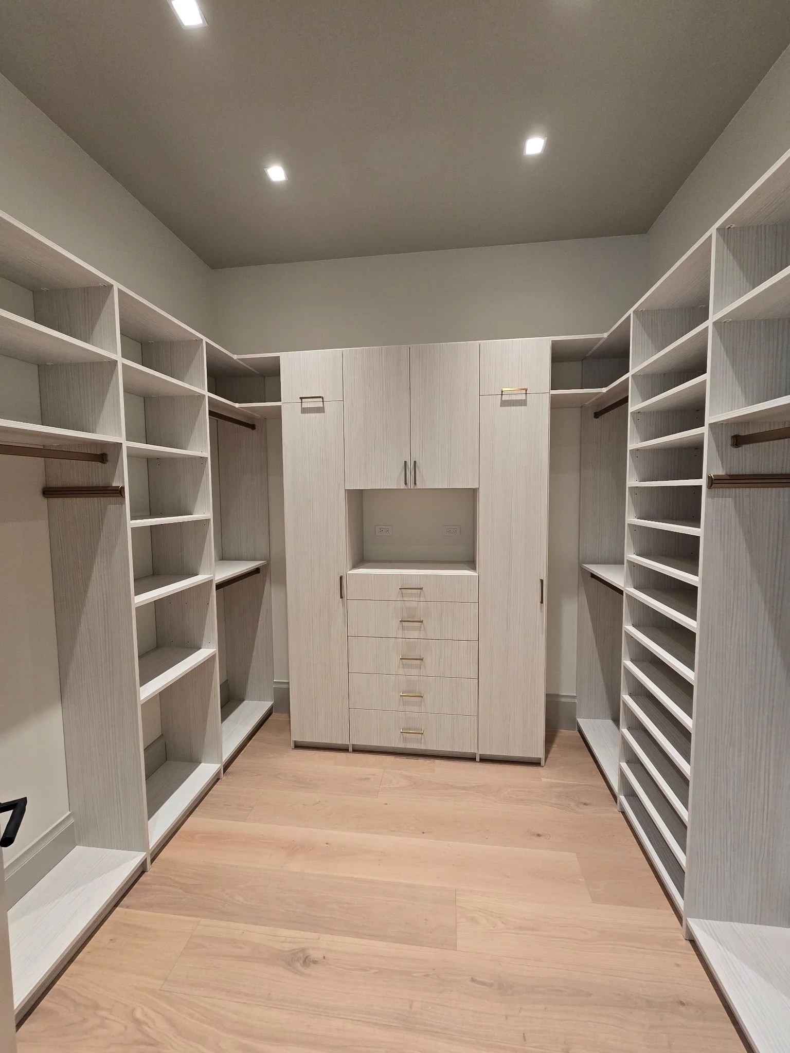 Empty walk-in closet with white built-in shelves, drawers, and hanging rods, and a wood-plank floor.