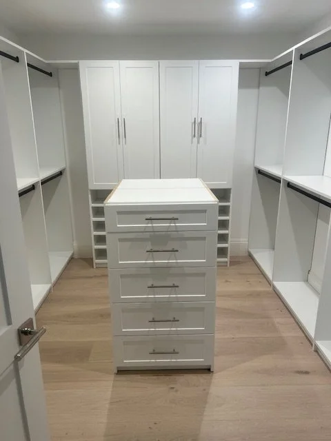 Empty walk-in closet with white built-in shelving and drawers, light wood flooring, and a central white storage island.