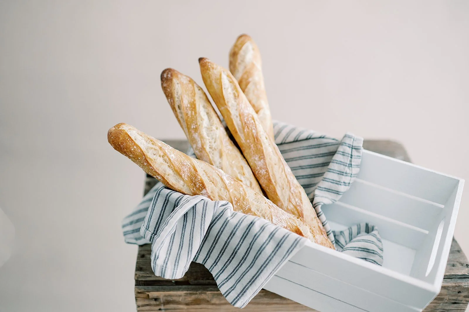 Several baguettes in a white basket lined with a striped cloth on a rustic wooden surface.