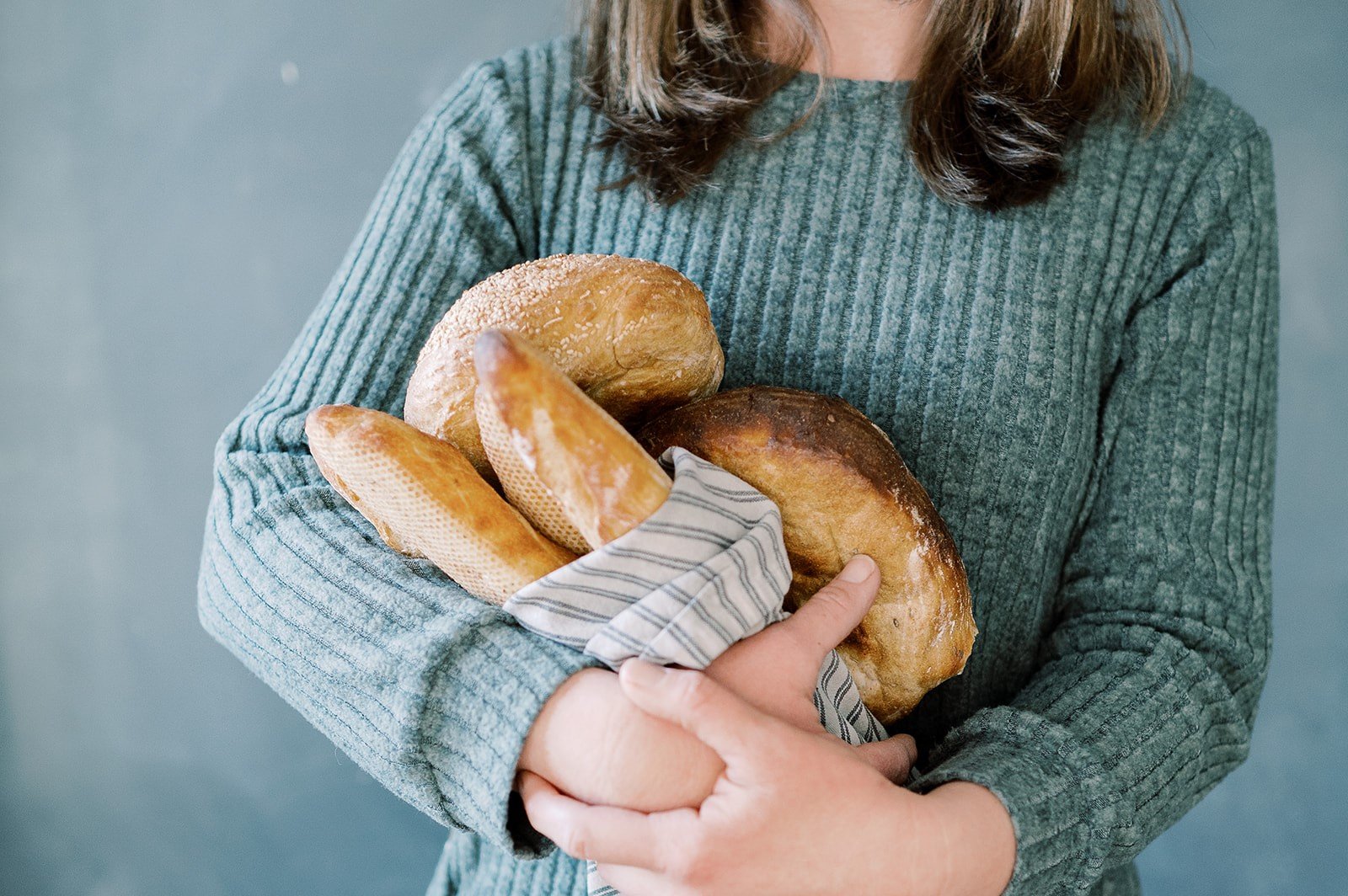 Person in a teal ribbed sweater holding a variety of bread loaves wrapped in a cloth.