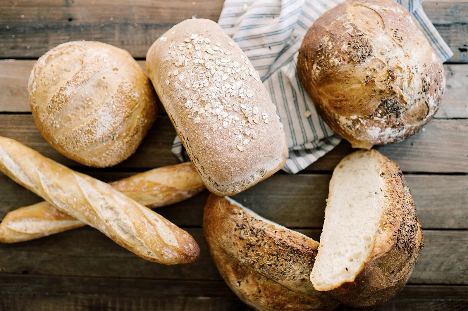 Assorted loaves of bread including baguettes, rolls, and sliced bread on a wooden surface.