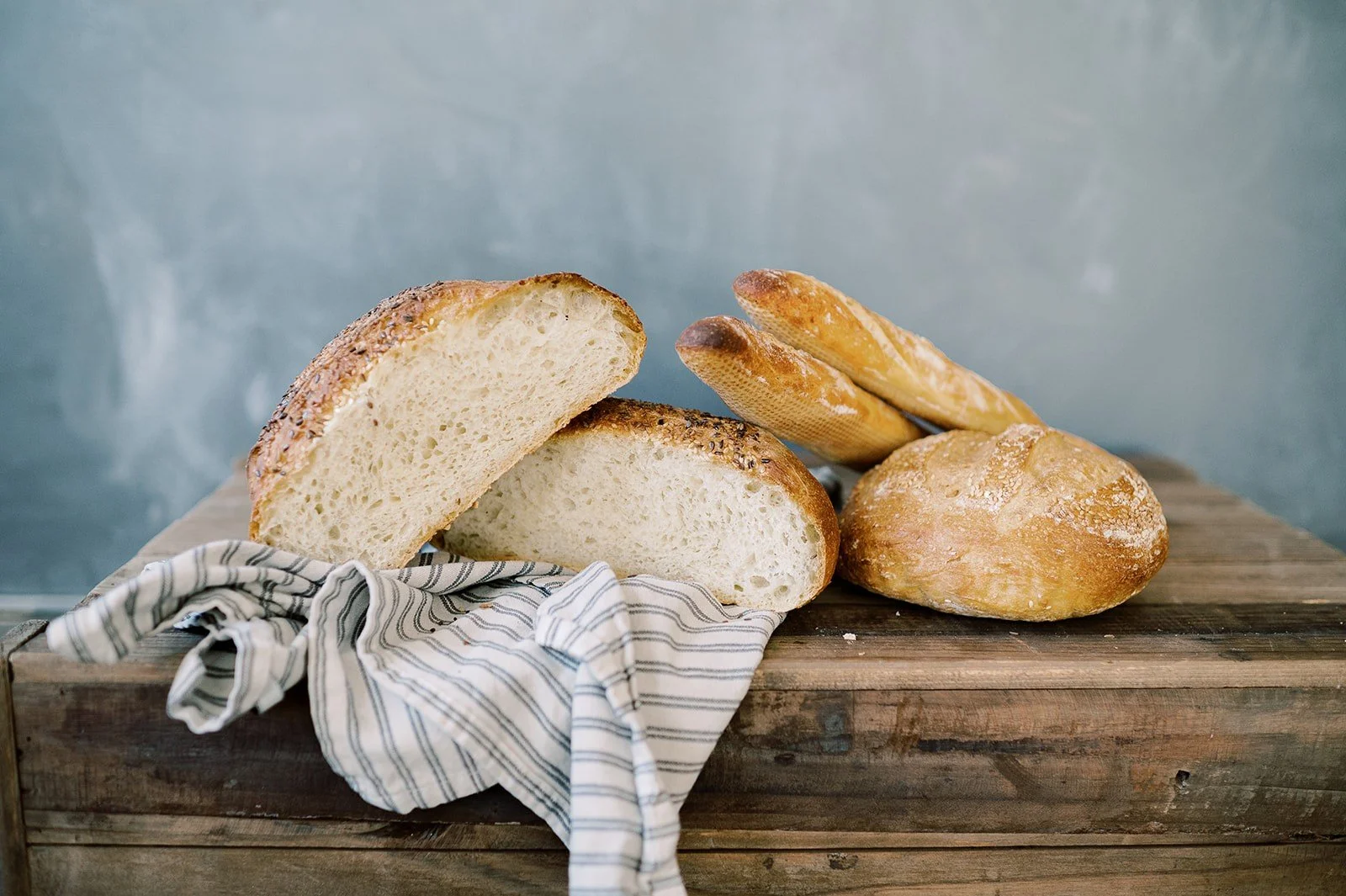 A loaf of sliced bread, a round bread roll, and two baguettes on a wooden cutting board, with a striped cloth underneath.