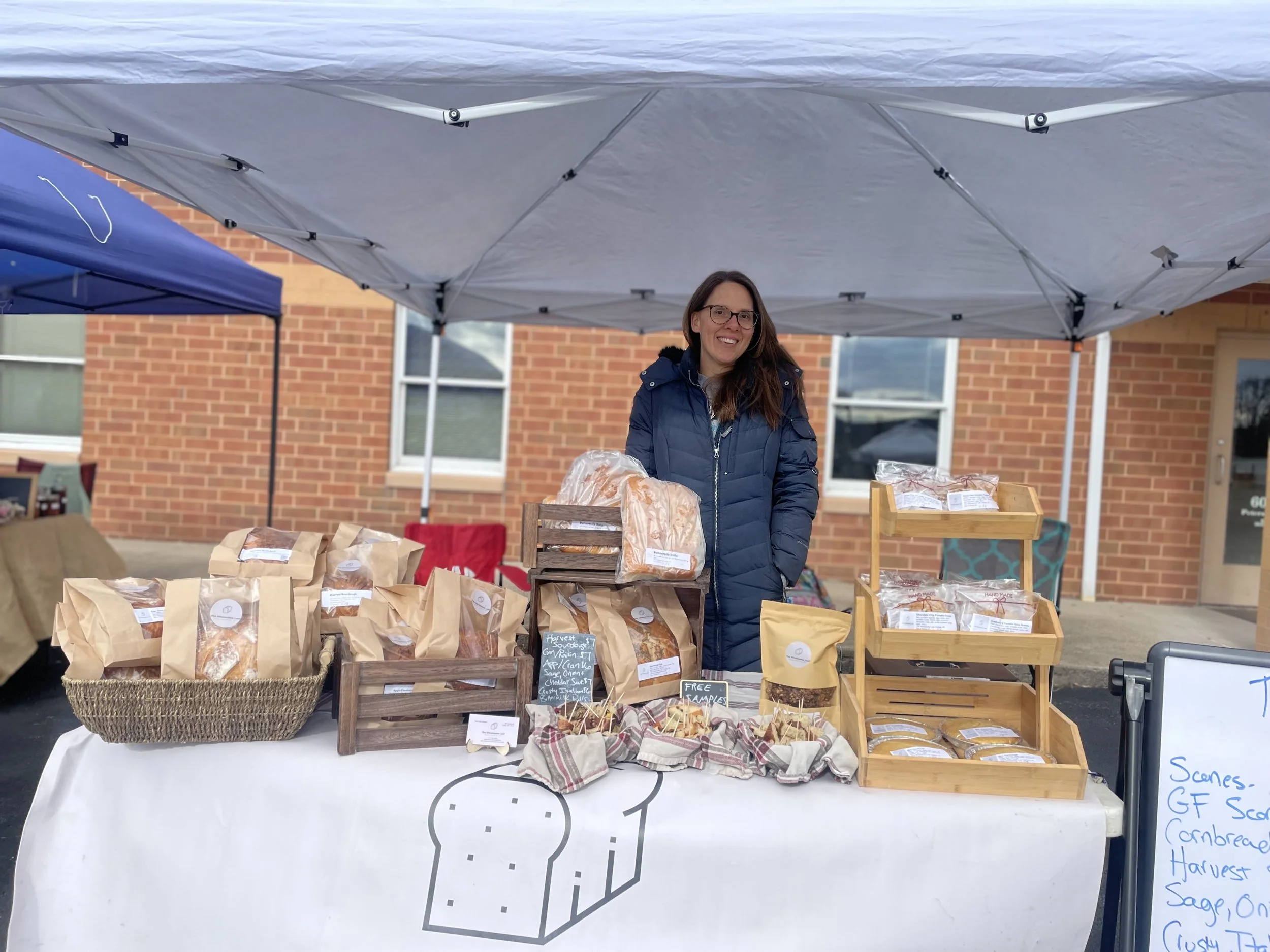 Woman selling baked goods at an outdoor market stall under a white canopy, with brick building in the background.
