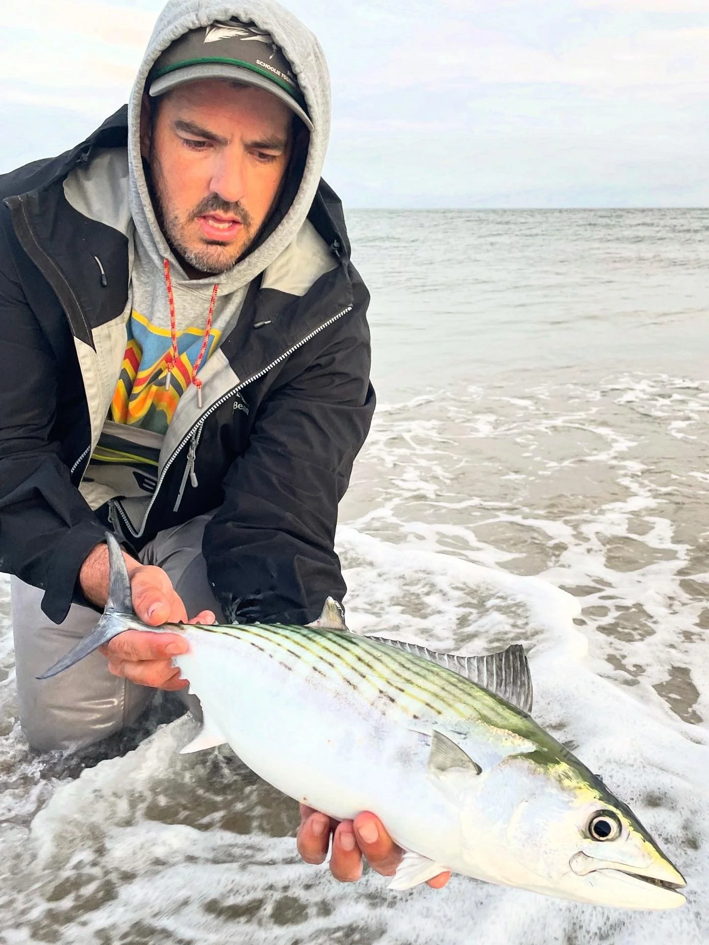 Man wearing a hoodie, jacket, and cap holding a large Bonito fish on the beach with the ocean in the background.