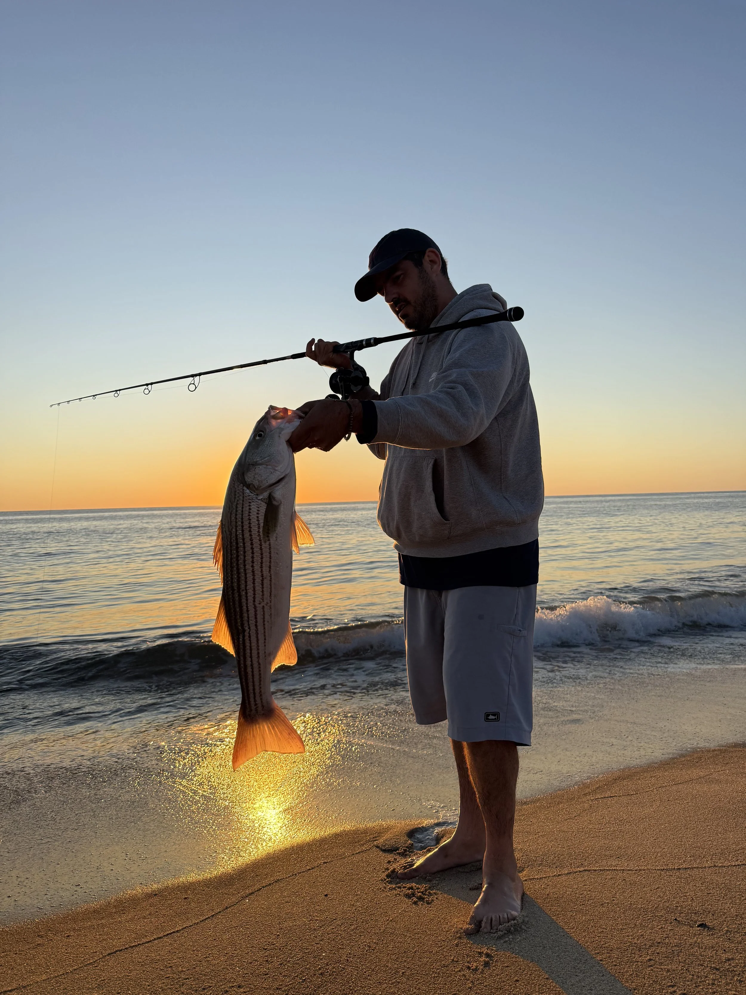 A man on the beach at sunset holding a fishing rod and displaying a large fish he caught.