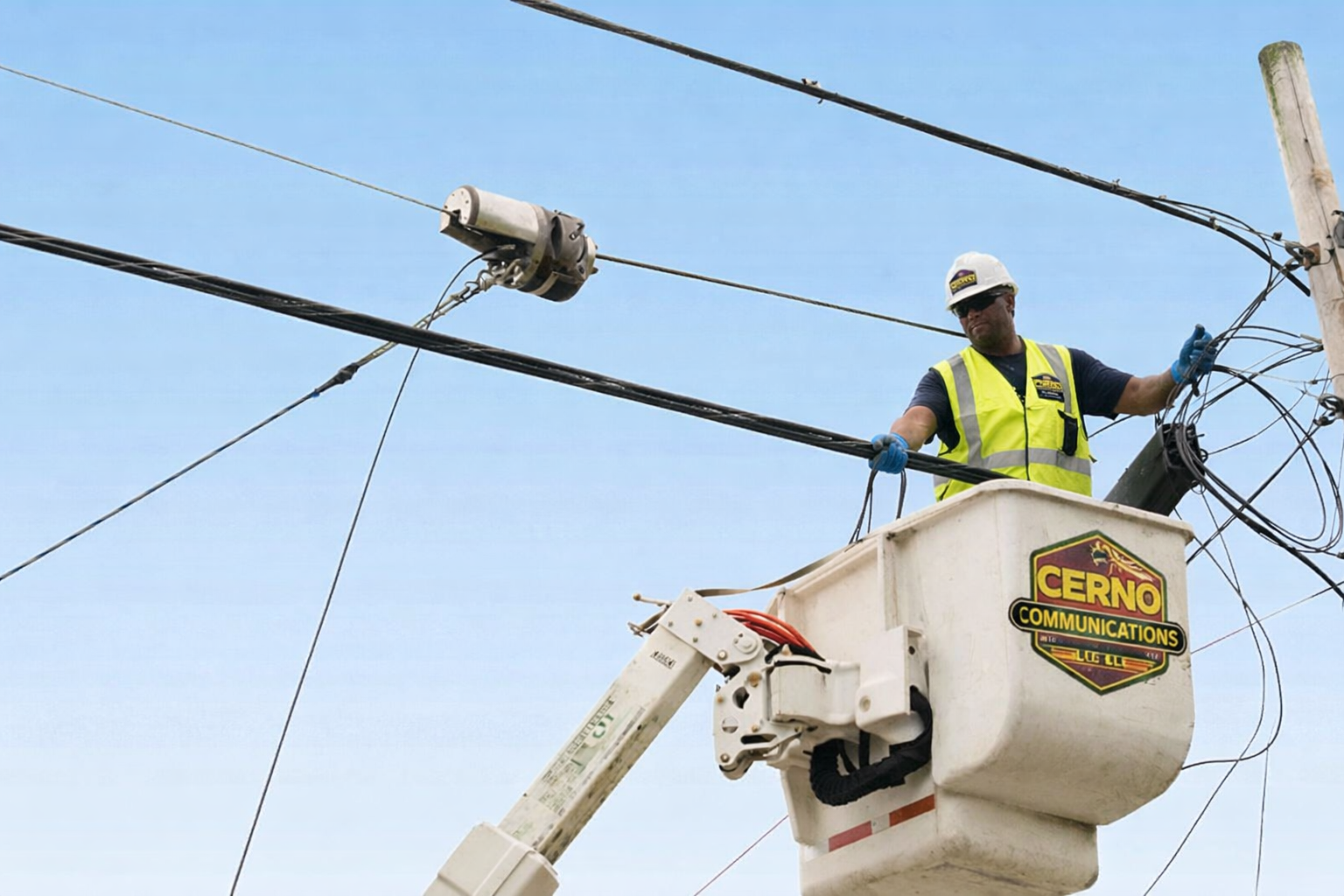 A utility worker in a yellow safety vest, helmet, and gloves standing in a bucket lift working on overhead power lines against a clear blue sky.